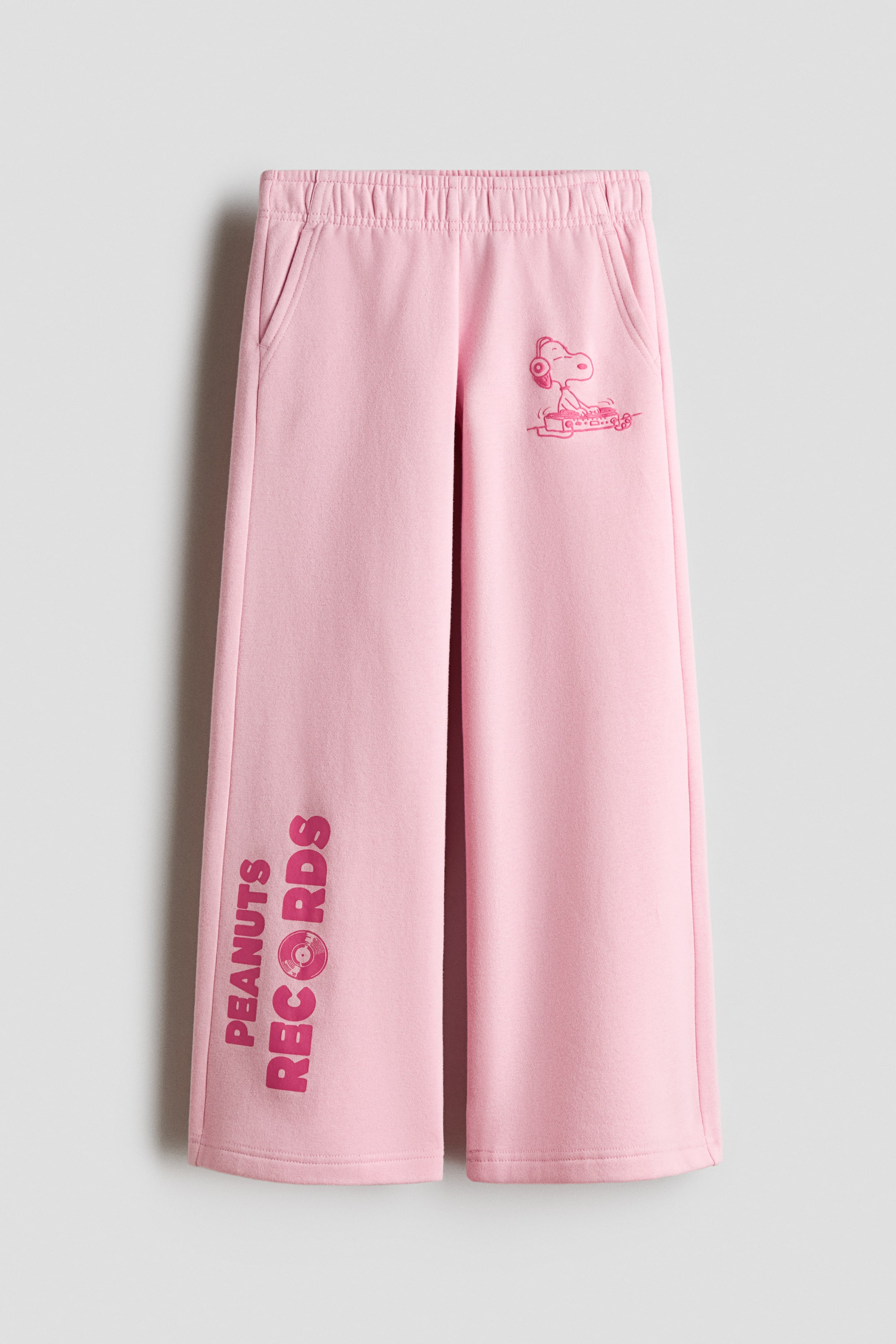 Grotere afbeelding bekijken: Een vooraanzicht van roze joggingbroek met een elastische tailleband, zijzakken en wijde, rechte pijpen. De broek toont een roze afbeelding van Snoopy met een koptelefoon op de linkerbovenbeen en verticale tekst "PEANUTS RECORDS" op het rechteronderbeen.