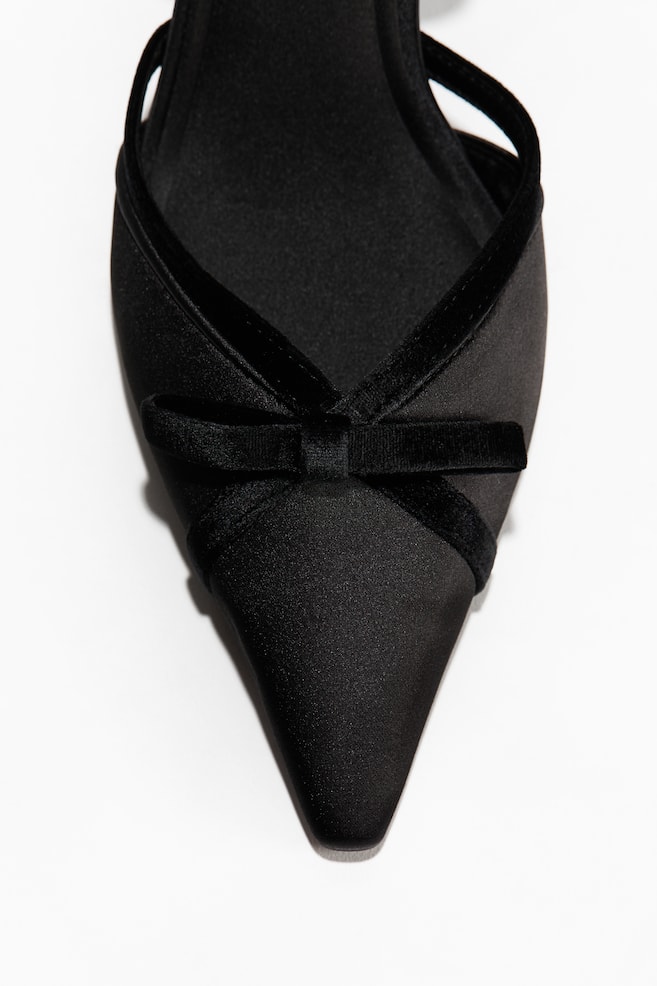 This close-up shot is showcasing a black satin pointed-toe shoe facing front. The shoe is featuring a black velvet trim along the collar that crosses over the vamp, where a small matching velvet bow is positioned. - 1