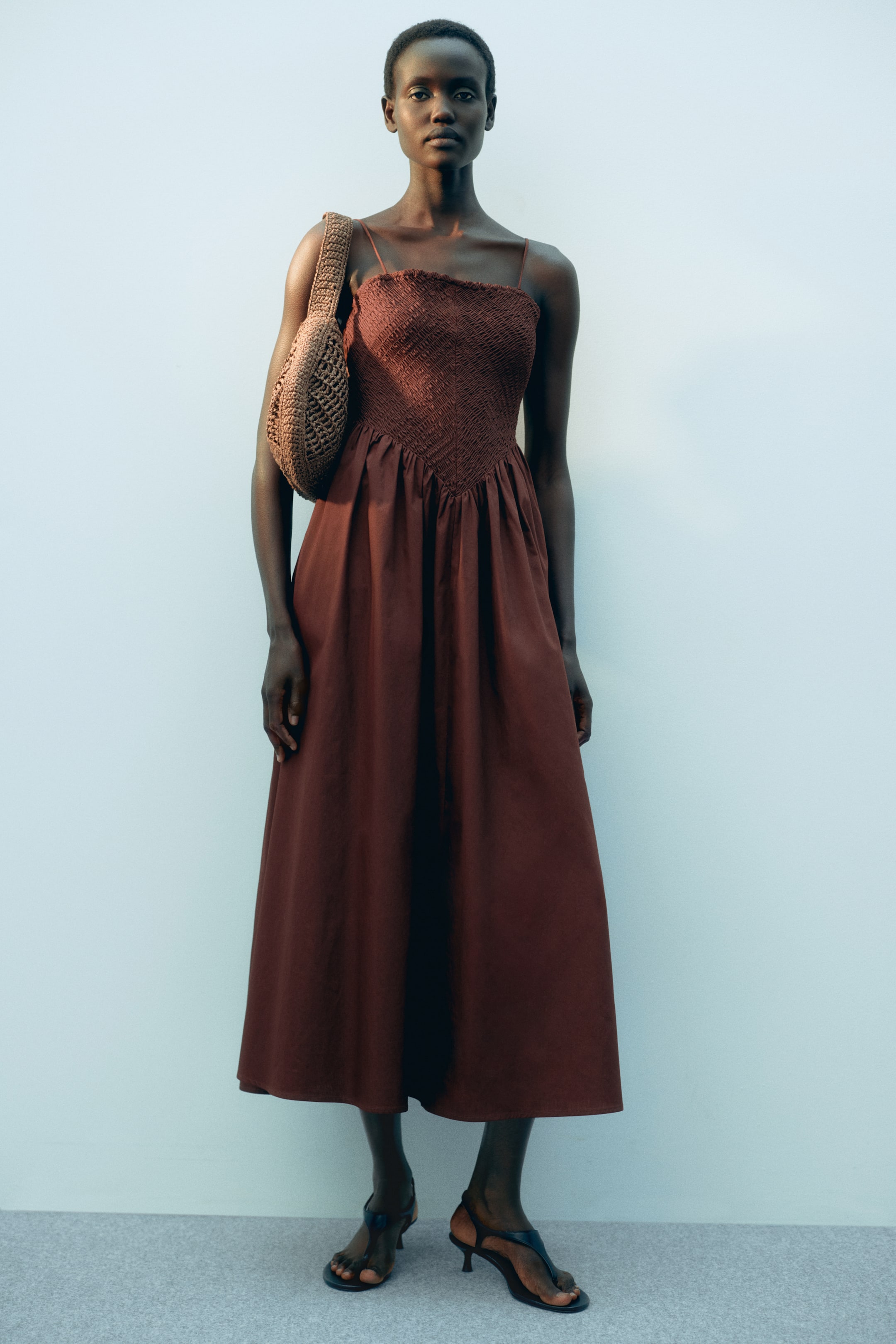 Ampliar la imagen: Un vestido marrón rojizo hasta la pantorrilla presenta un corpiño fruncido con escote recto y tirantes estrechos; una costura fruncida en forma de V define la cintura y desde ella se desprende una falda acampanada. La modelo lleva el vestido, porta un bolso tejido al hombro y sandalias negras.