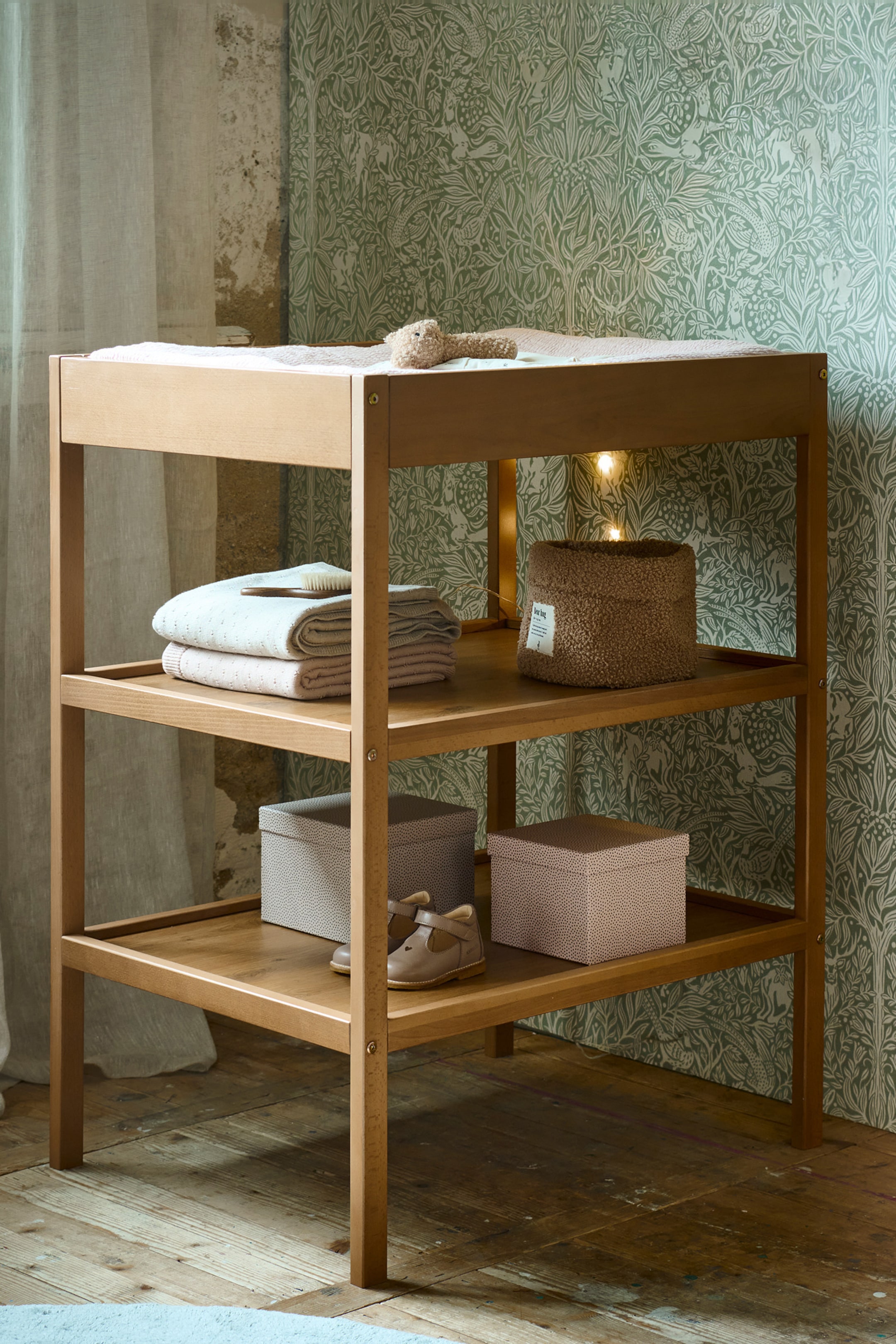 Grotere afbeelding bekijken: Een drielaagse commode van licht hout is iets naar links gekanteld, waardoor de voorkant en rechterkant zichtbaar zijn. Op de bovenste plank ligt een lichtgekleurde verschoningsmat met een klein knuffeldier, en op de middelste plank liggen gevouwen lichtgekleurde textiel en een bruine, gestructureerde mand. De onderste plank bevat twee opbergdozen met patroon en een paar babyschoentjes.