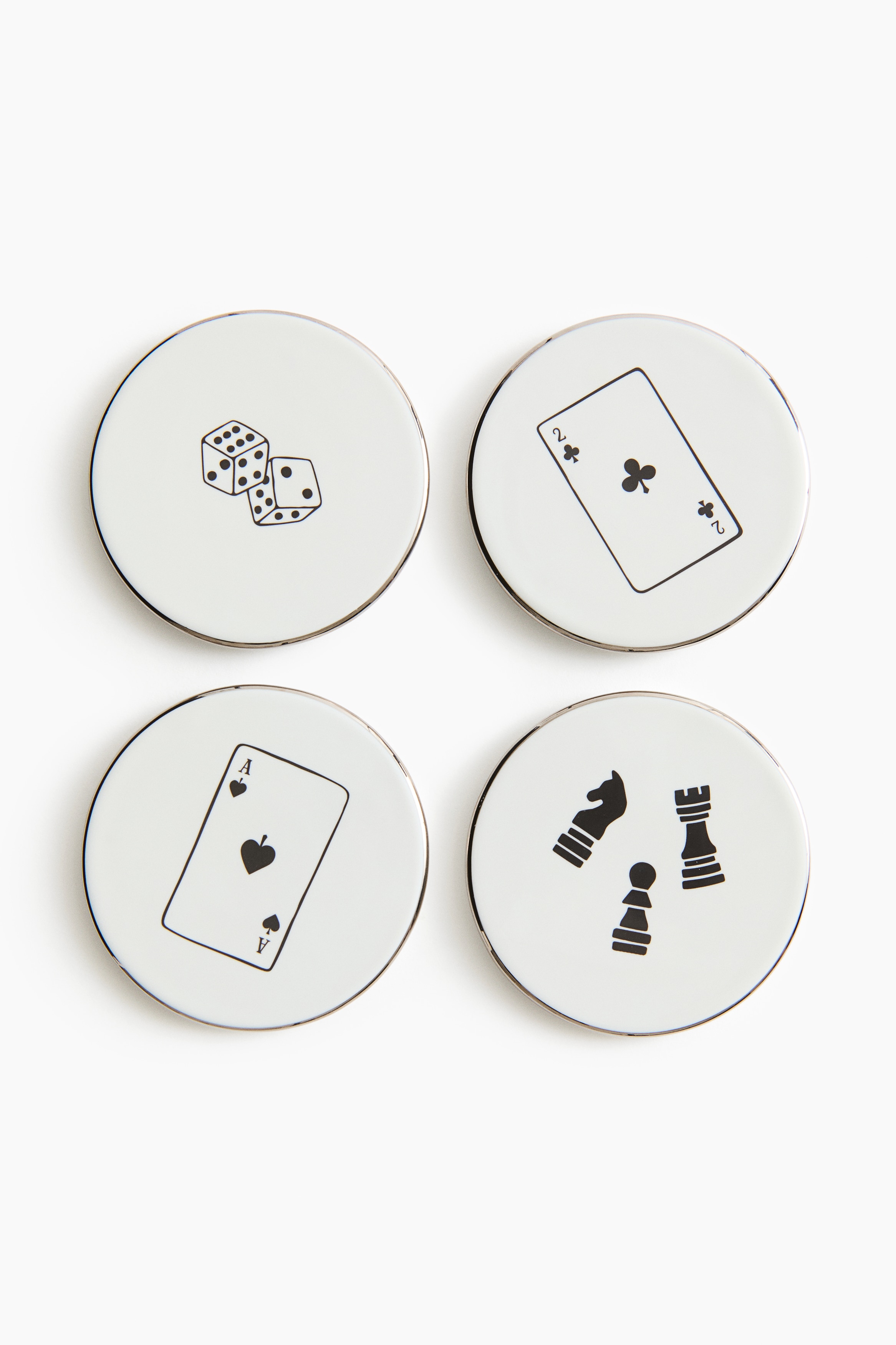 Four circular porcelain coasters are displayed in a top-down view, each featuring unique black graphic designs. Three of the coasters show illustrations of an Ace of Spades card, a pair of dice, and a Two of Clubs card, while the fourth coaster supports a clear glass filled with liquid.