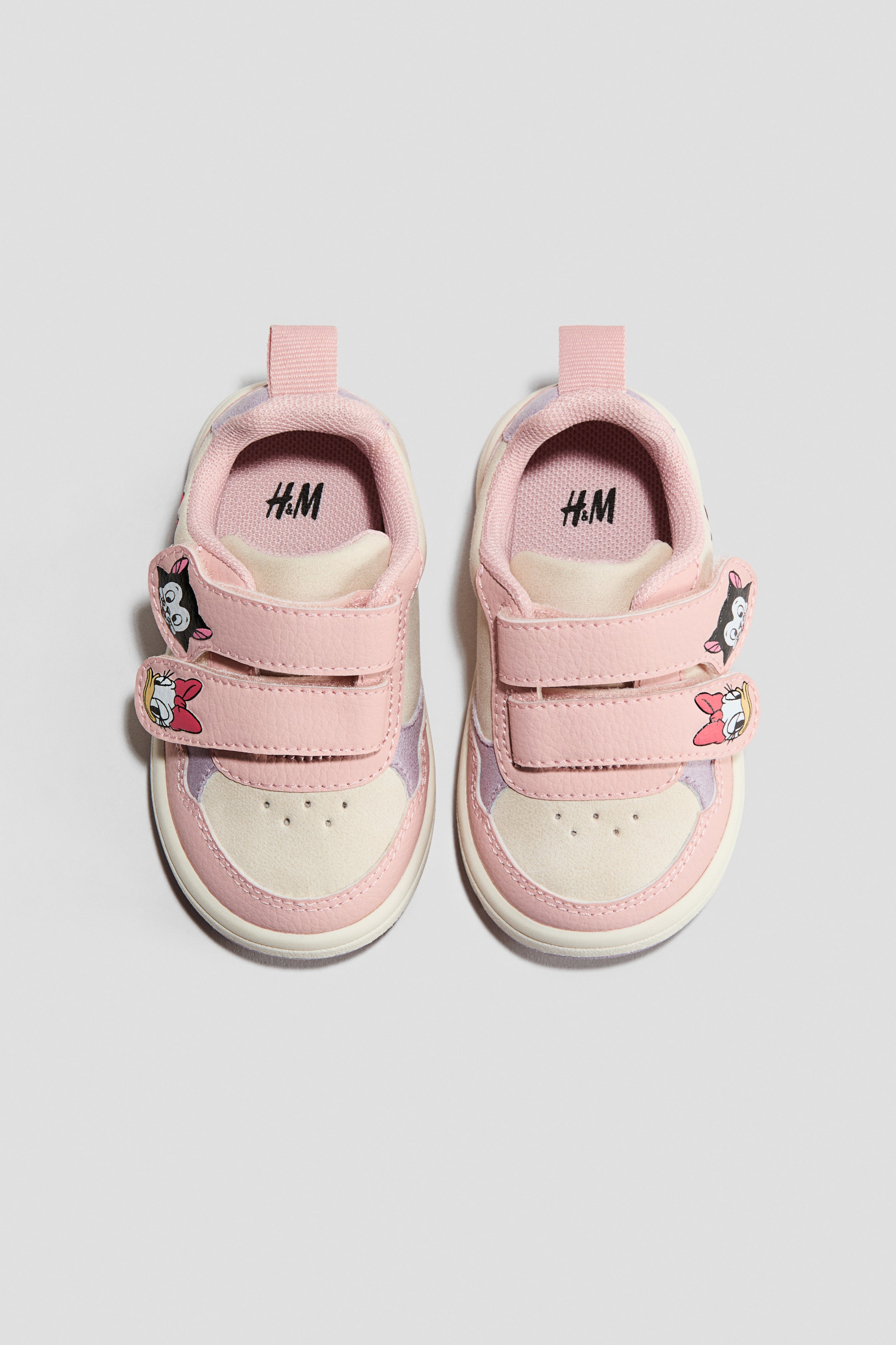 Ampliar la imagen: Un par de zapatillas para bebé se presenta como FRONT, con un esquema de colores crema y rosa pálido con acentos en púrpura claro. Cada zapato está asegurado con dos correas de velcro, que están adornadas con apliques de personajes de dibujos animados.