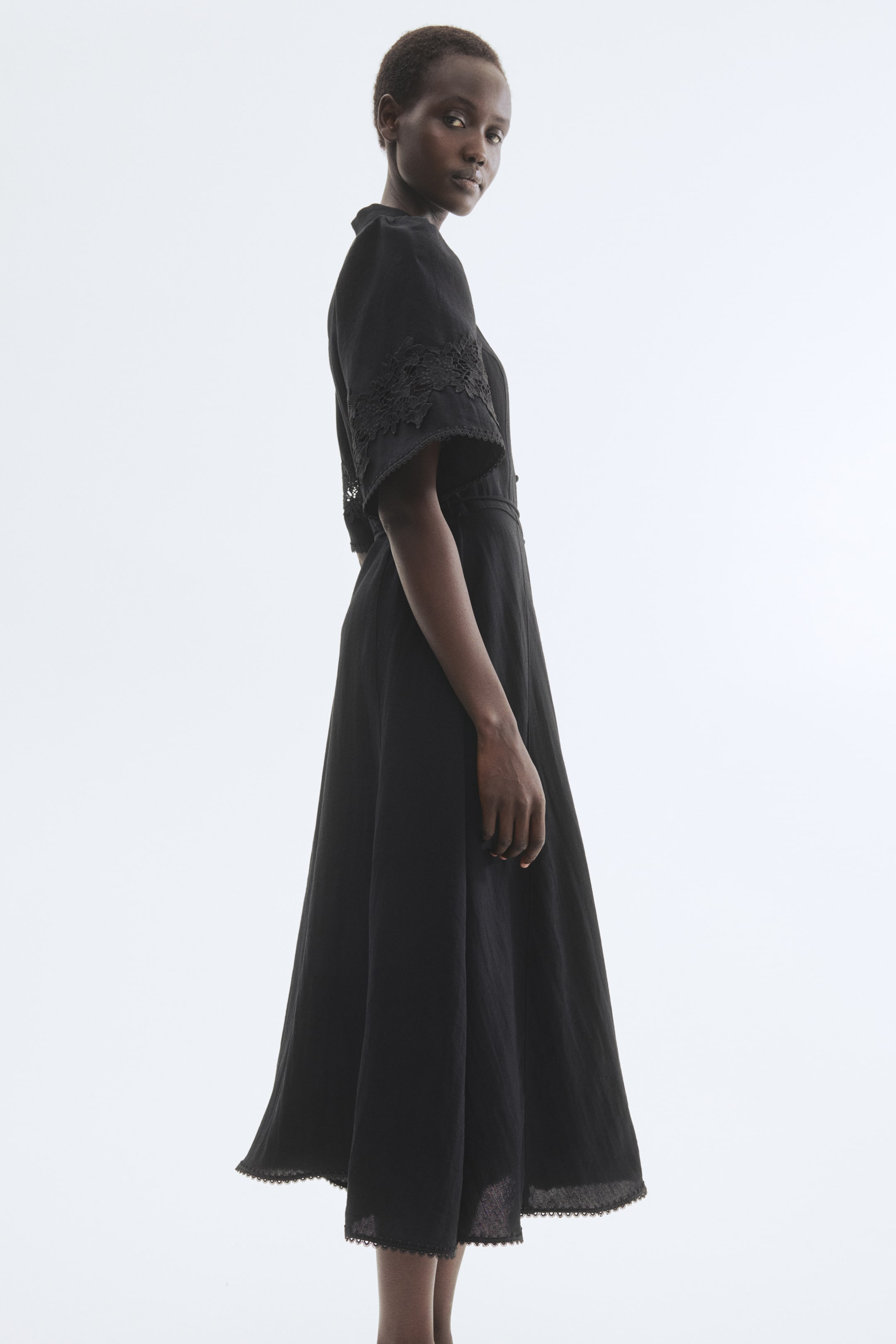 View larger image: "A black midi dress is shown from a side-back angle, featuring a high neckline and short, wide sleeves. The sleeves are detailed with floral lace appliqué and scalloped trim, which is also appearing along the flared skirt's hem."