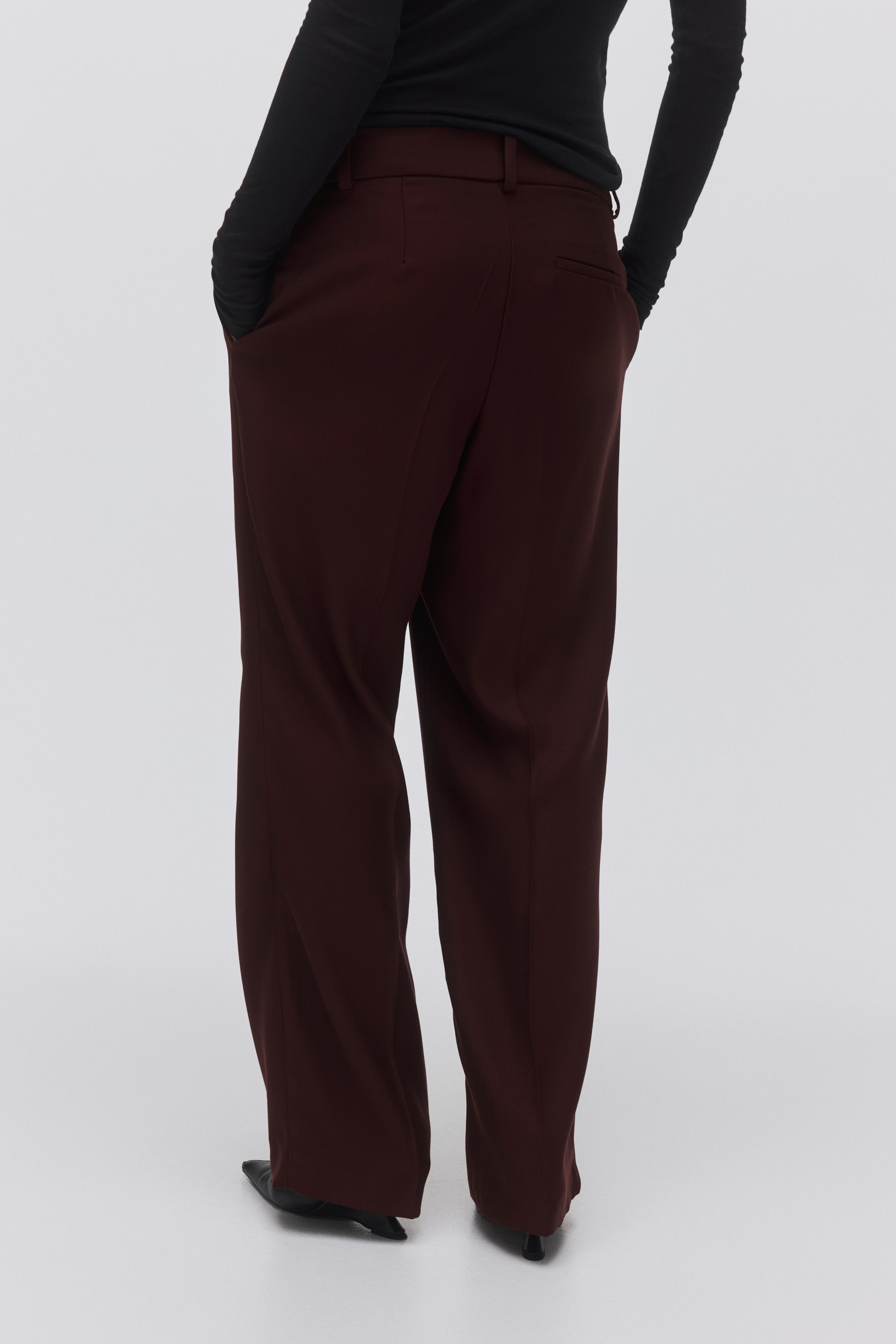 Agrandir l'image: Un pantalon large de couleur bordeaux est présenté sous un angle arrière, avec des passants de ceinture et une poche arrière passepoilée. Le pantalon présente une coupe ample et large, et les mains reposent dans les poches latérales.