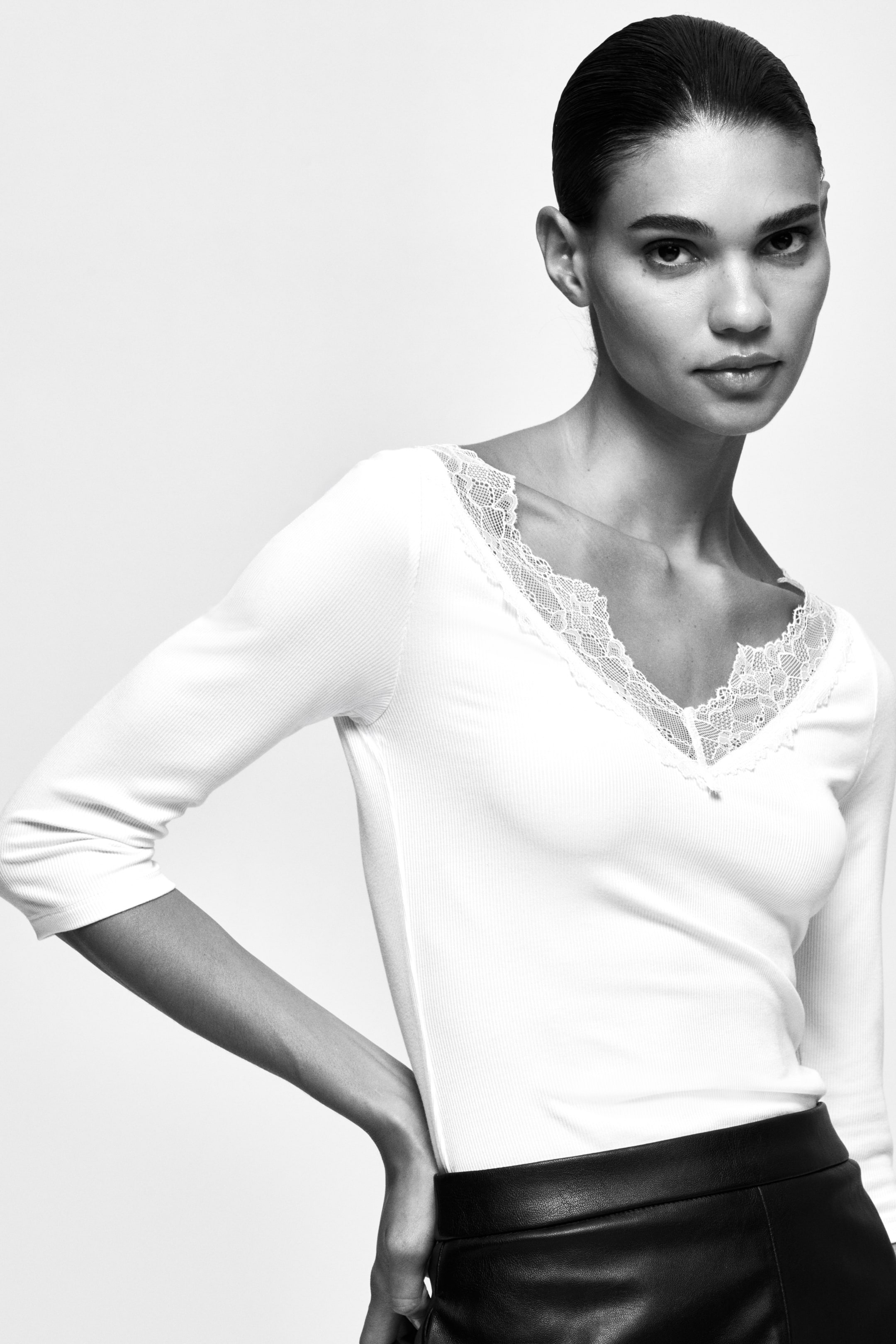View larger image: The model is wearing a white ribbed top with a V-neck and lace trim. It has three-quarter sleeves and is paired with a black high-waisted skirt.