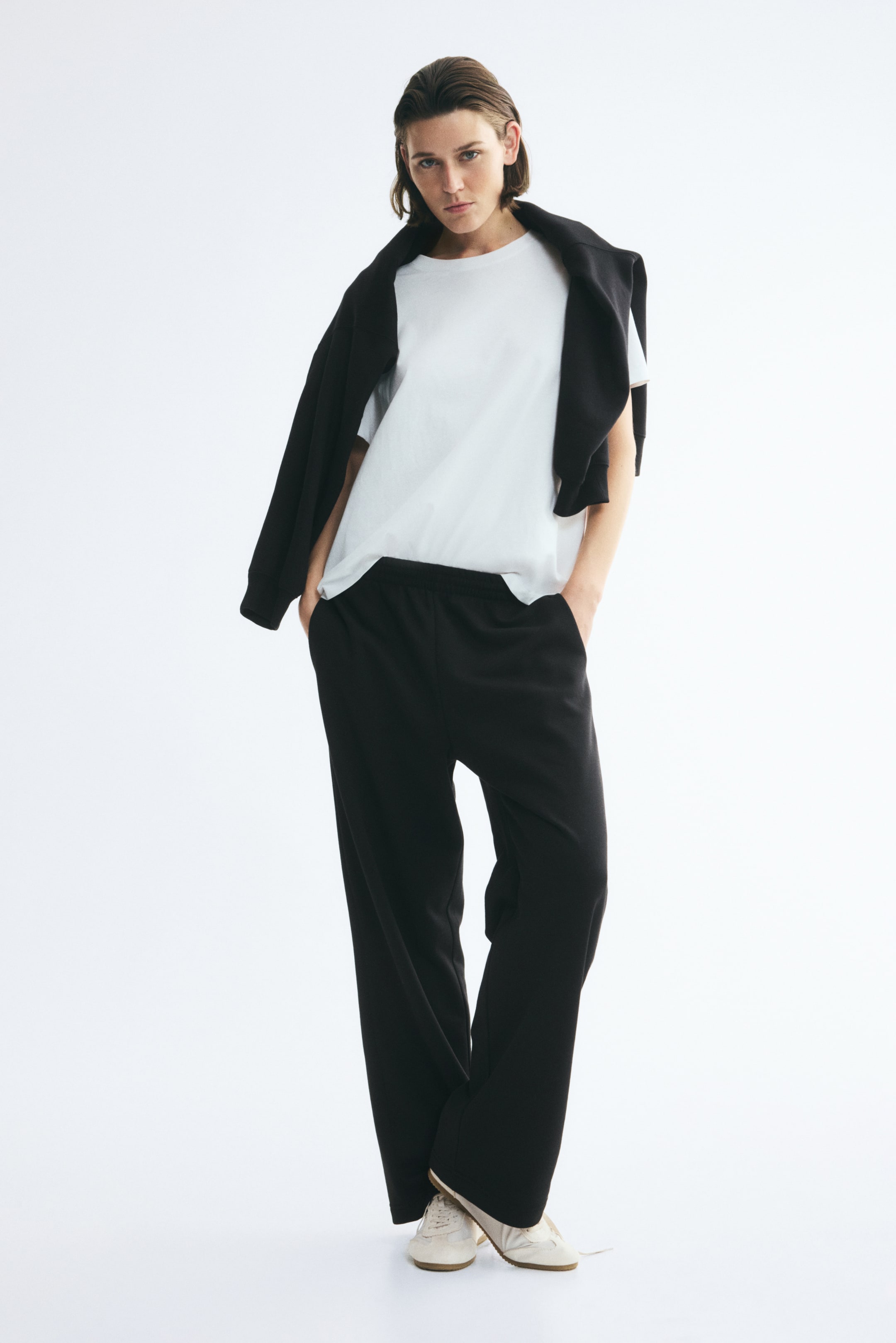 View larger image: Black track pants featuring straight legs and an elasticised waistband are being worn by the model, who is standing with hands placed in the side pockets. The pants are styled with a white t-shirt, a black sweatshirt draped over the shoulders, and light-coloured sneakers.
