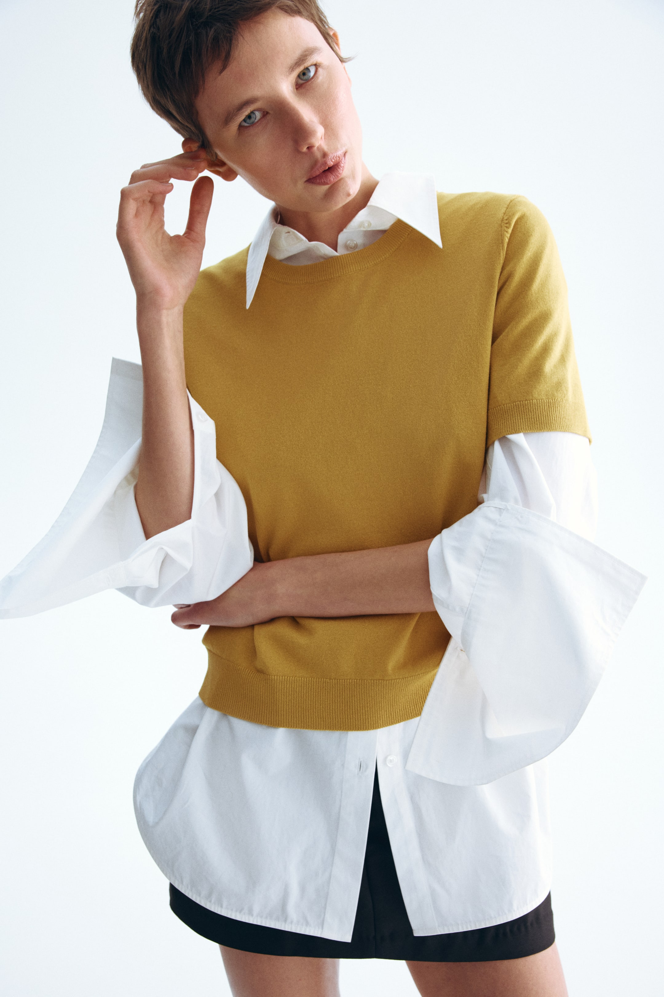 View larger image: A mustard yellow short-sleeved sweater with a crew neck and ribbed hem is layered over a white collared shirt. The shirt's pointed collar is visible above the sweater, its long bell sleeves extend from beneath the sweater's short sleeves, and its curved hem is visible below the sweater.