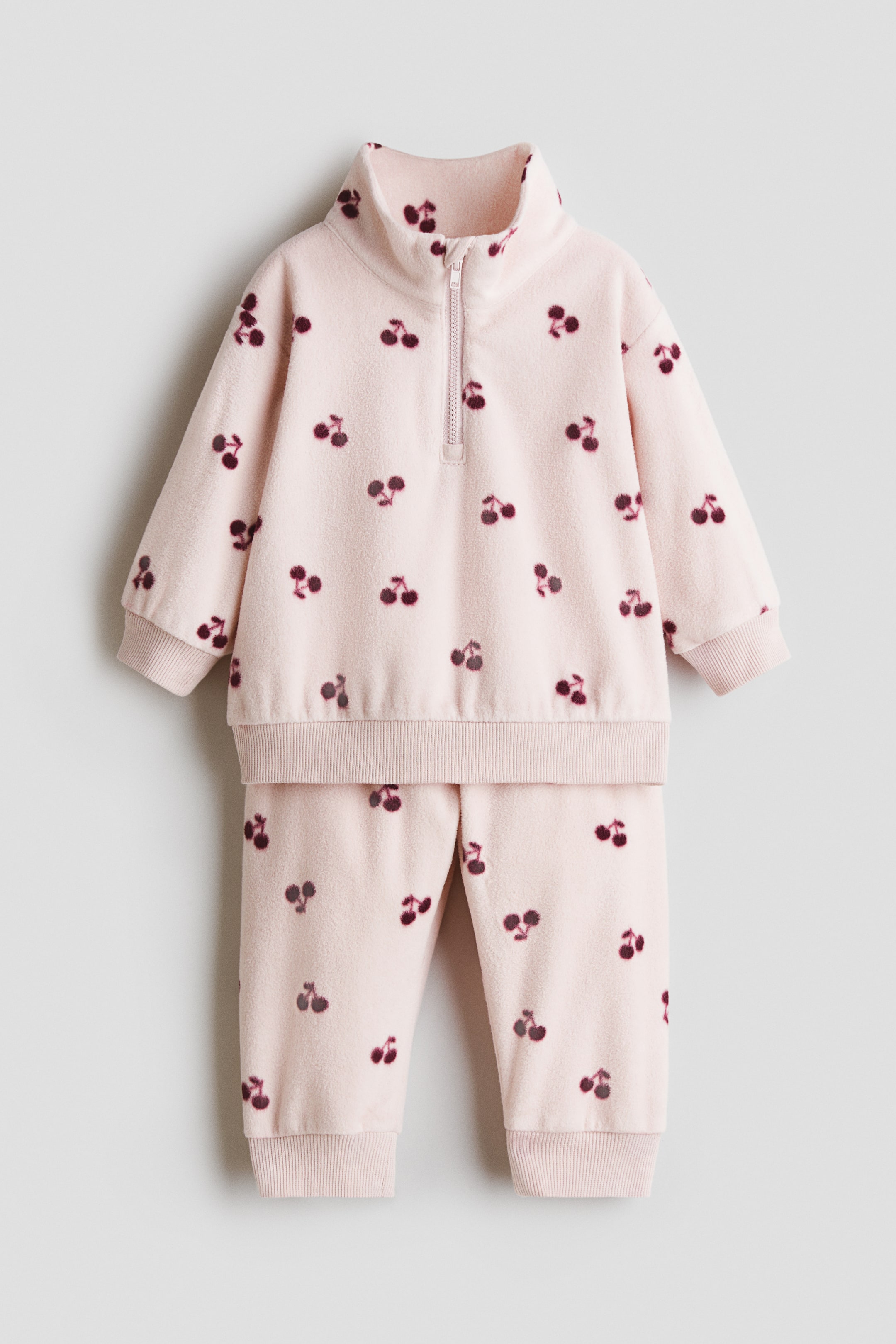 View larger image: A light pink two-piece baby outfit is shown from the front, featuring a pullover top and matching pants. Both pieces showcase an all-over dark cherry pattern, with the top featuring a stand-up collar, a half-zip closure, ribbed cuffs, and a ribbed hem, while the pants feature ribbed ankle cuffs.