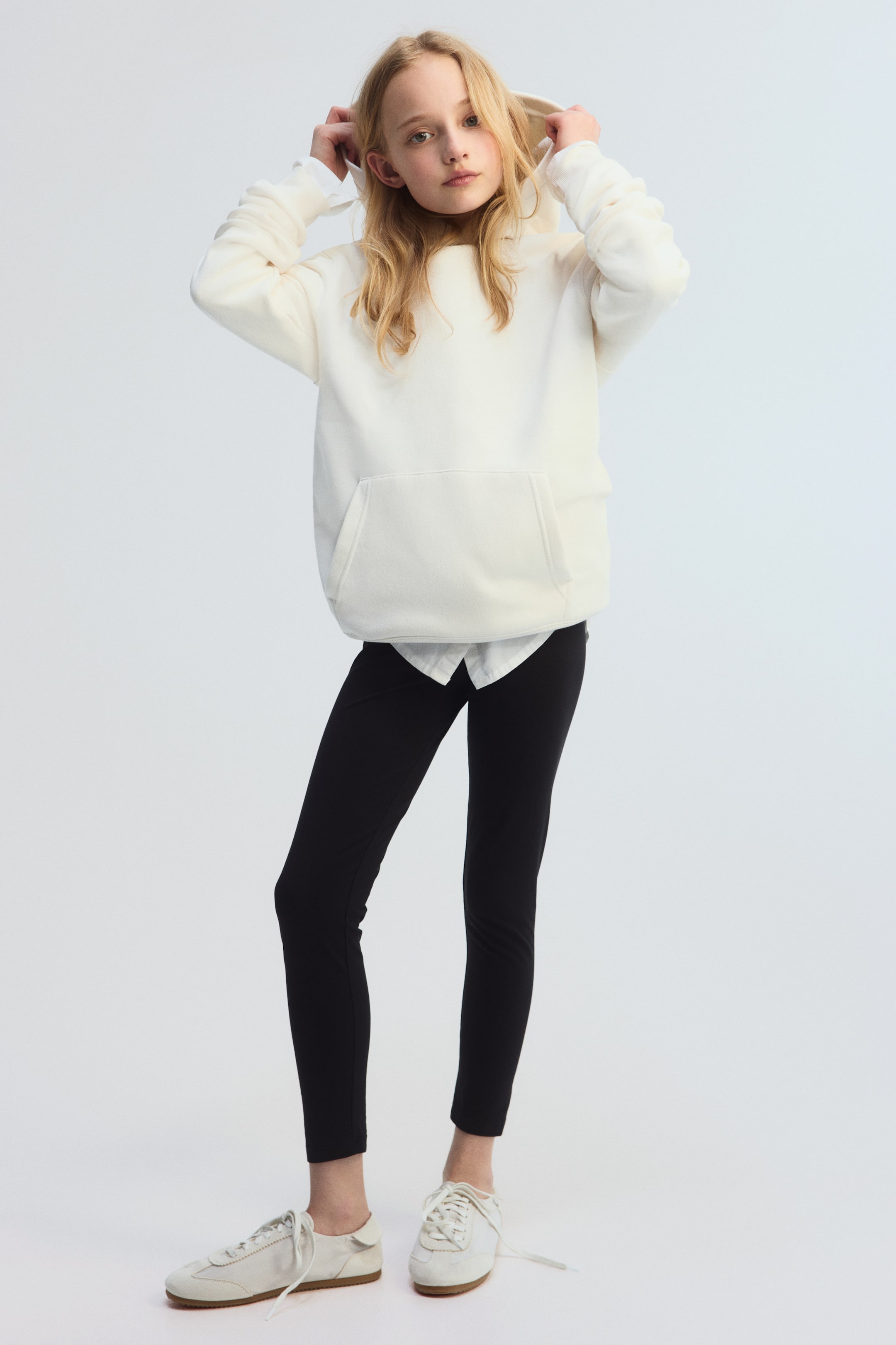 View larger image: An off-white oversized hoodie is showcased from a slight front-left angle, featuring a soft, fleece-like texture and a large kangaroo pocket. A hood is visible, with a white collared shirt peeking out from beneath the hoodie's hem.