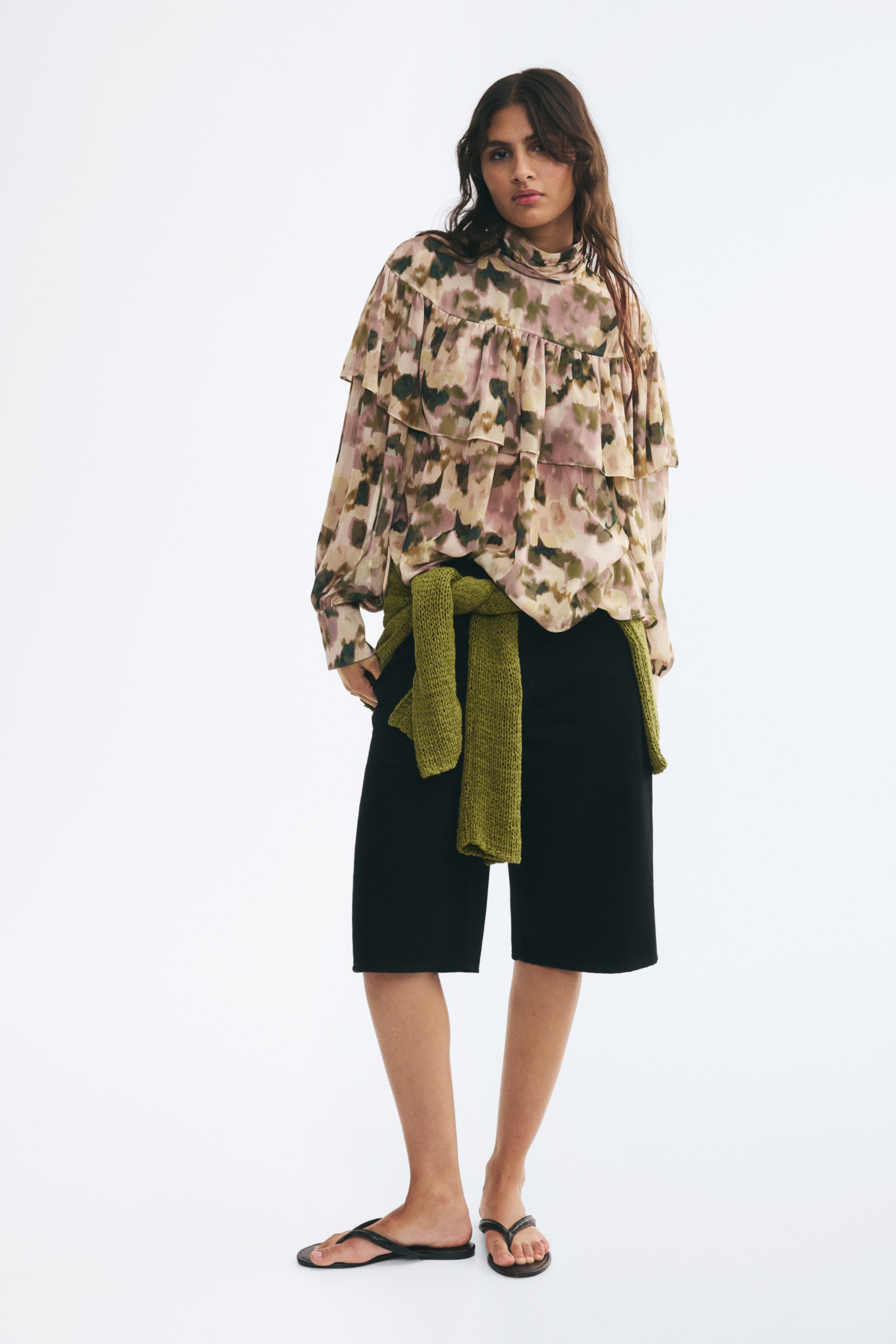 View larger image: An oversized, semi-sheer blouse featuring a watercolour-style pattern in muted pink, green, and brown tones is worn by the model. The blouse features a high neck, a ruffled flounce yoke, and long balloon sleeves, and is styled with black knee-length shorts and a green knit sweater tied around the waist.