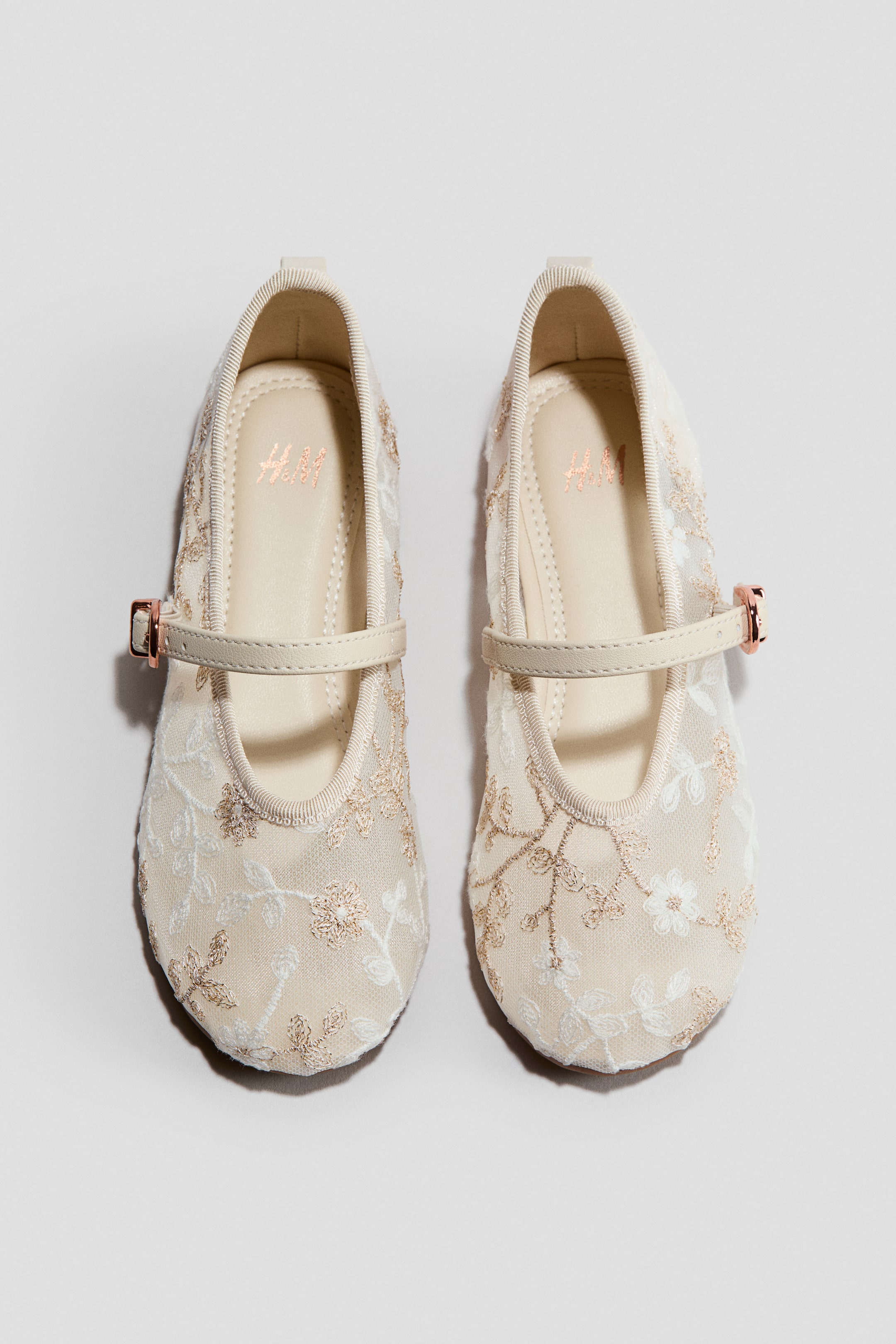 Visa större bild: Ett par krämfärgade ballerinaskor har en genomskinlig meshovandel, intrikat broderad med vita och guldfärgade blomsterdetaljer. Varje sko har ett krämfärgat band över vristen, fäst med ett roséguldfärgat spänne och en ljus inläggssula med en märkeslogotyp.