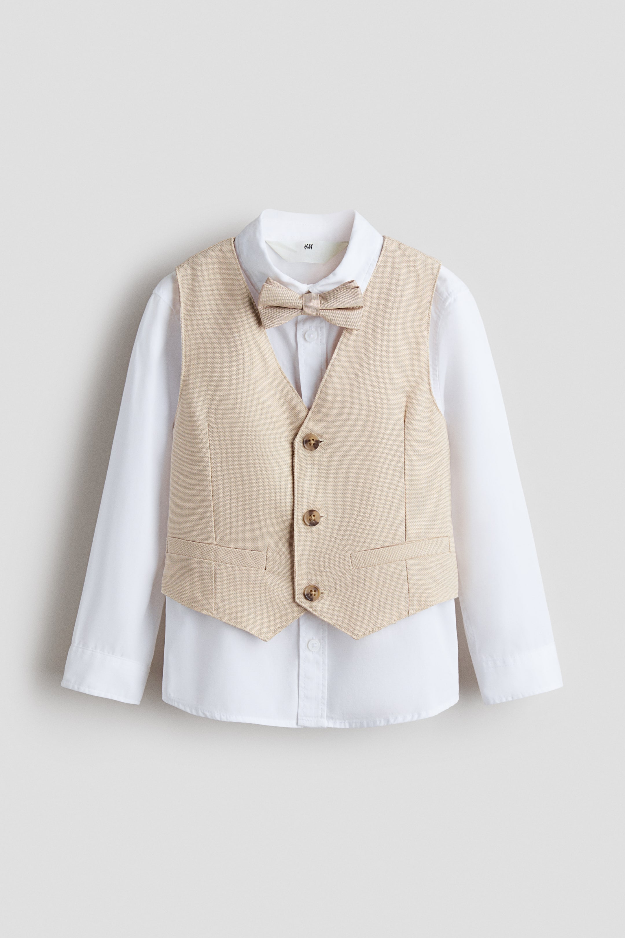 Größeres Bild ansehen: Ein schickes Set wird VORNE gezeigt, mit einem weißen Hemd, einer beige gewebten Weste und einer passenden vorgebundenen Fliege. Das Hemd präsentiert einen Kragen, Knöpfe entlang der Vorderseite und lange Ärmel mit geknöpften Manschetten, während die Weste einen V-Ausschnitt, drei Vorderknöpfe und zwei falsche Taschen zeigt.