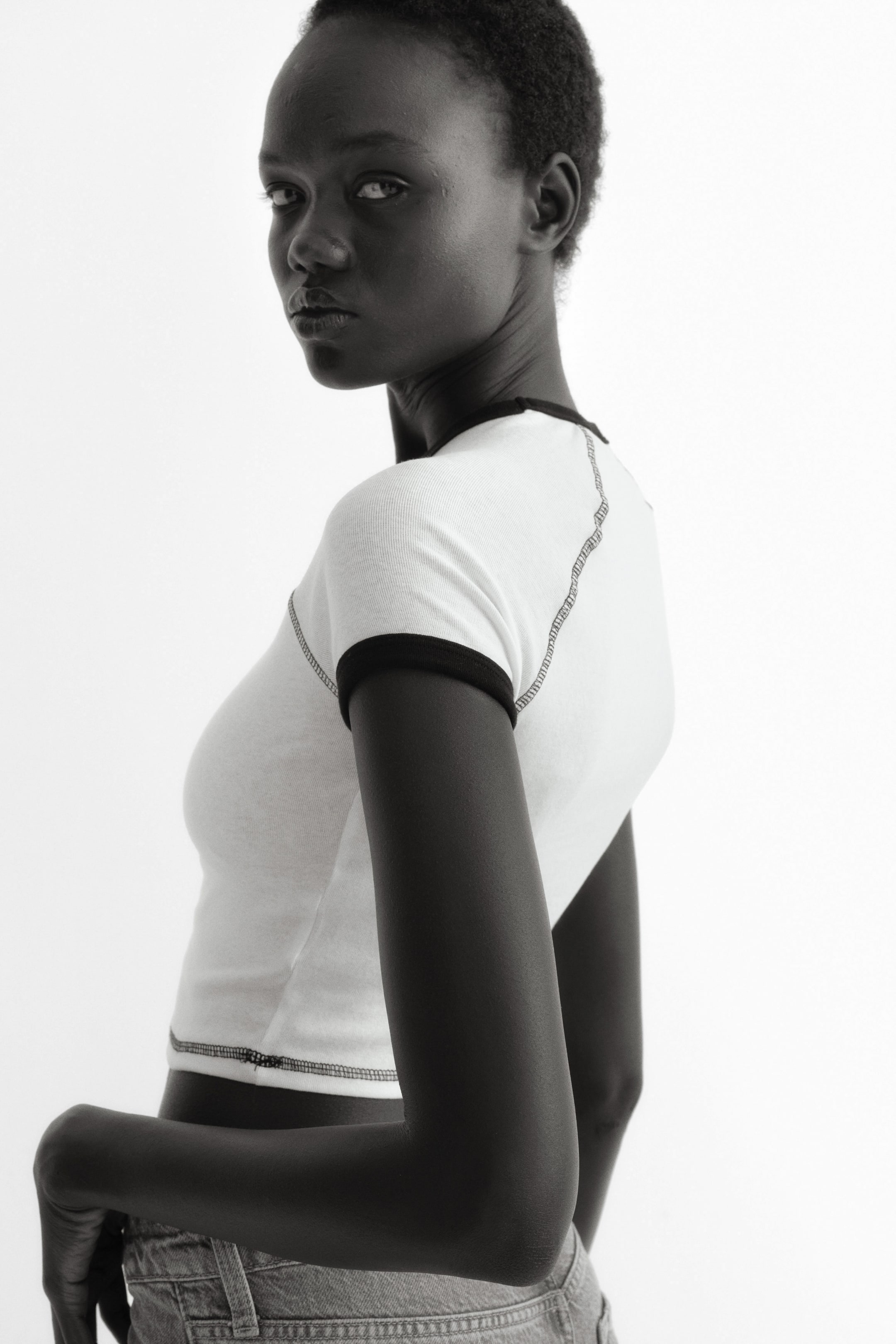 View larger image: A white ribbed ringer T-shirt featuring black contrast trim on the crew neck and short sleeves, shown from a slightly angled back view. Black exposed stitching highlights the shoulder seams, side seams, and bottom hem, and the top is designed in a cropped length.