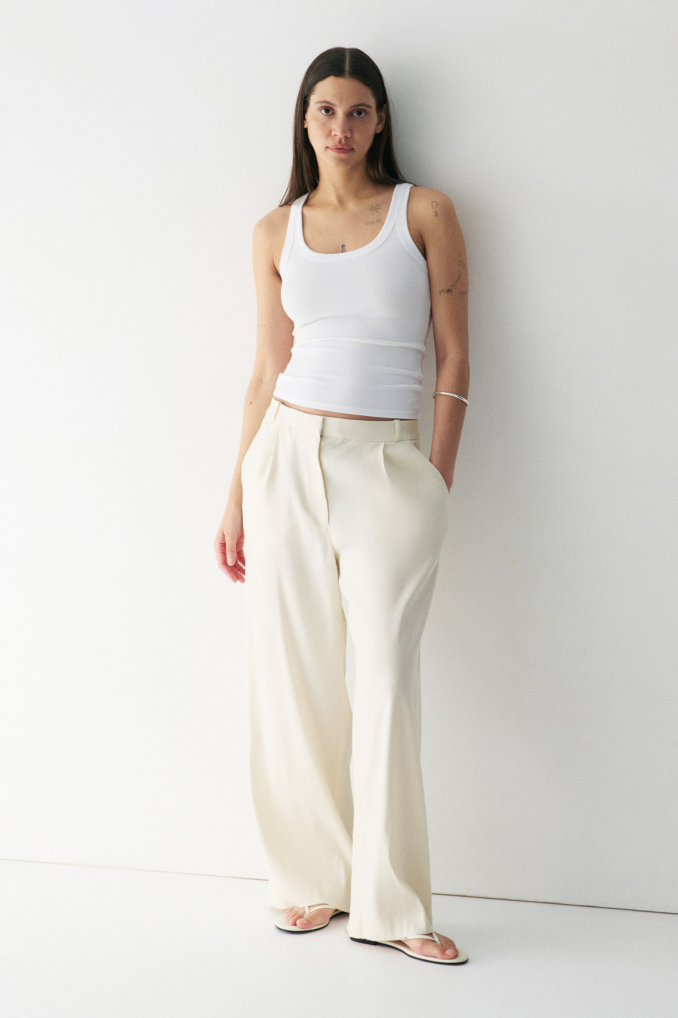 View larger image: A white ribbed vest top is being worn, featuring a scoop neck, broad straps, and a form-fitting silhouette, ending just above the waistband. Cream wide-leg trousers are being worn, featuring a flat front, single pleats, and side pockets, all viewed from a slight front-left angle.