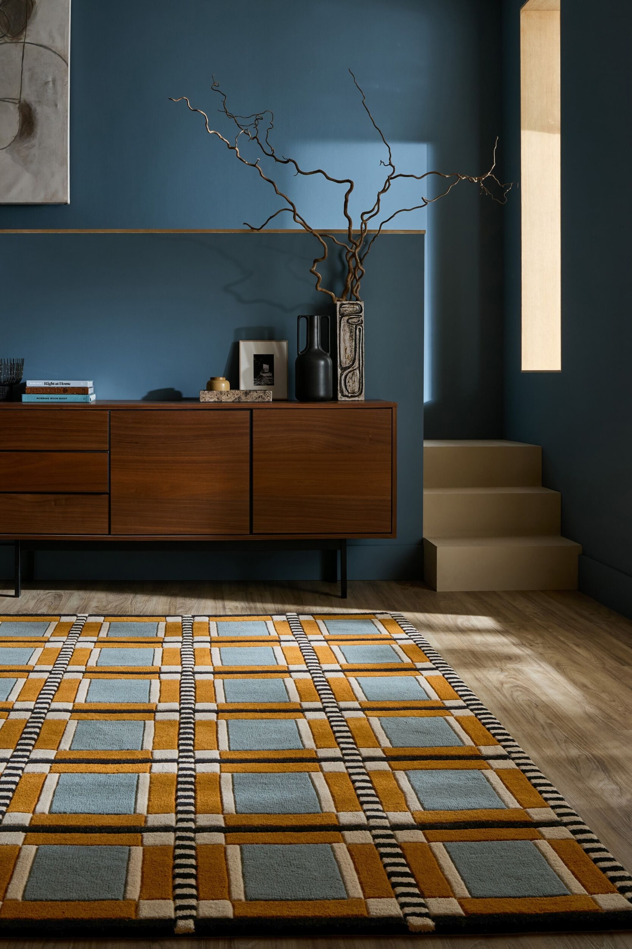 Ver imagen más grande: La alfombra presenta un patrón geométrico de cuadrícula en amarillo mostaza, azul claro, crema y negro, con grandes cuadrados azul claro enmarcados por rectángulos en amarillo mostaza y crema, y unidos por franjas texturizadas verticales y horizontales de rayas alternas en negro y crema.