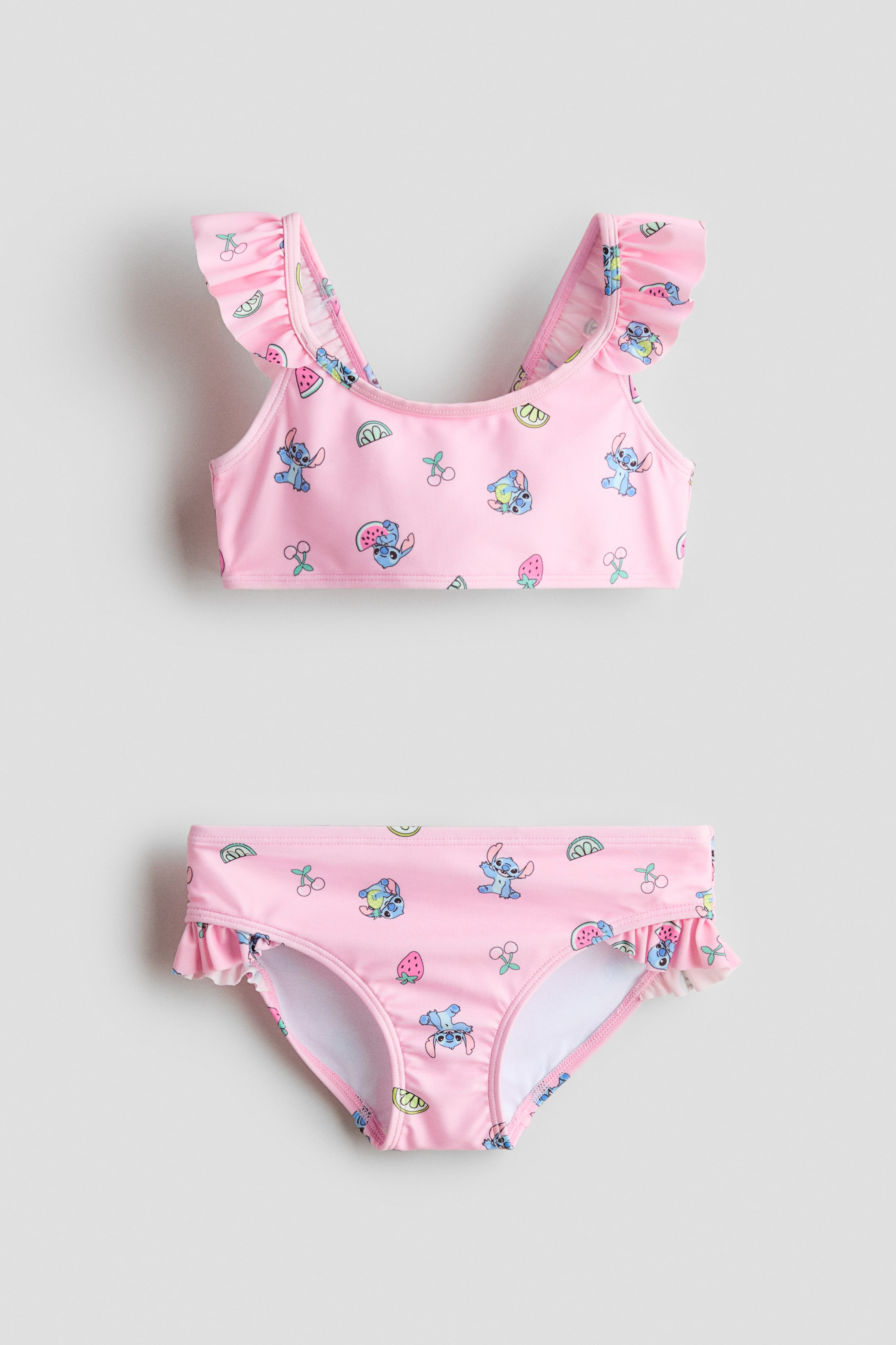 View larger image: A fully lined, pink bikini set is showcased at the front, featuring a print of Stitch characters and fruits. The top is designed with a round neckline and flounced shoulder straps, while the matching bottoms feature a flounce trim at the top of the leg openings.