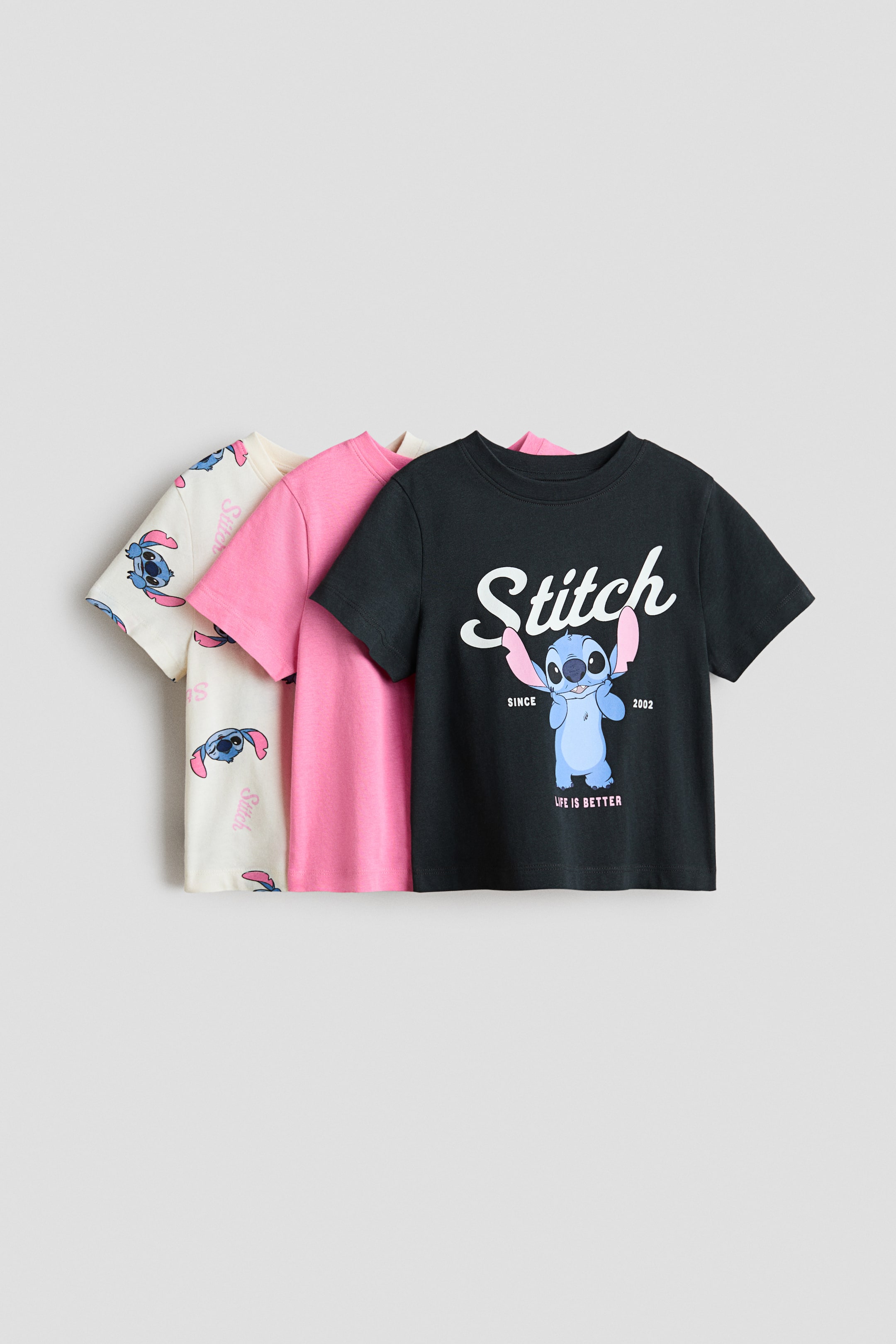 Xem ảnh lớn hơn: Ba chiếc áo thun ngắn tay với họa tiết in đang được xếp hàng, trình bày mặt trước của chúng. Chiếc áo đen ở phía trước nhất có hình nhân vật Stitch lớn và chữ, tiếp theo là chiếc áo màu hồng trơn, và chiếc áo màu trắng ngà có họa tiết nhân vật Stitch và chữ lặp lại.