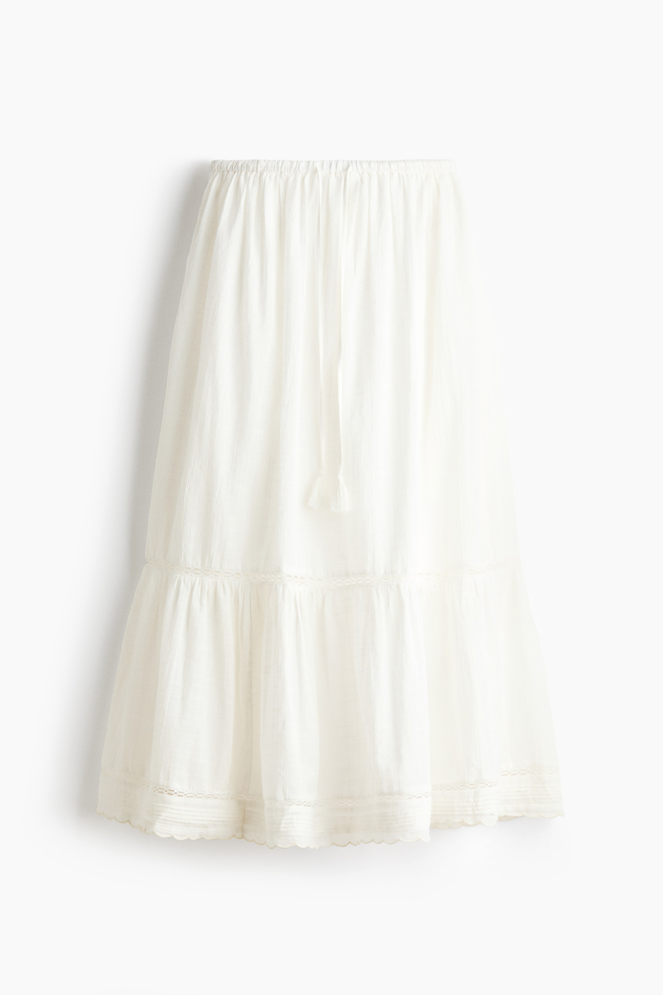 View larger image: The white tiered skirt is facing front. It features an elasticated waist with a drawstring and tassels, showcasing a lace trim along the tiered seam and a scalloped hem with added lace detailing.