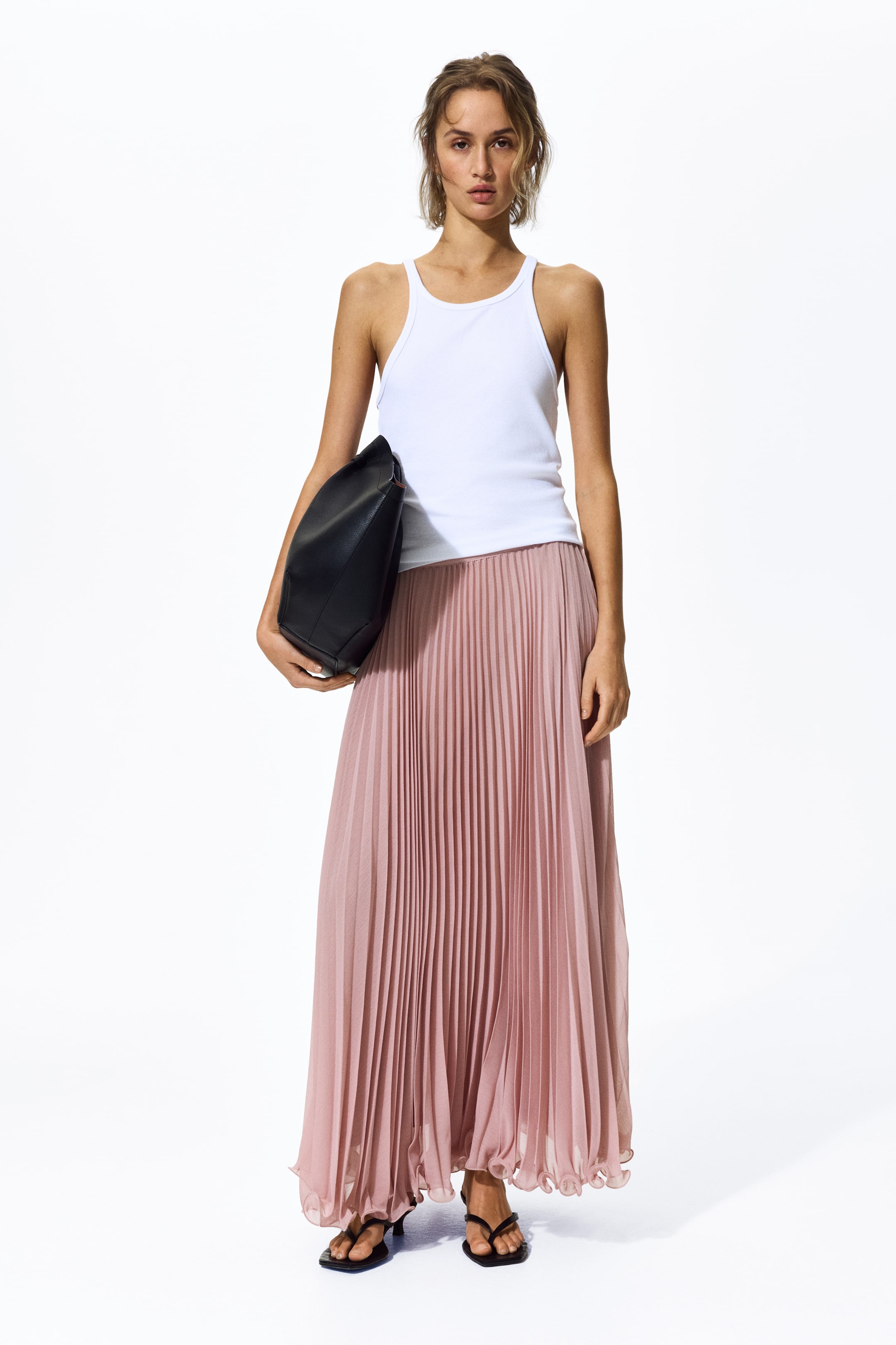 Grösseres Bild ansehen: Ein weißes, geripptes Tanktop mit Rundhalsausschnitt und breiten Trägern wird getragen, in einen langen, hellrosa plissierten Maxirock gesteckt. Eine schwarze, lässige Tasche wird an der Seite gehalten, und schwarze, eckige Zehensandalen runden das Outfit ab.