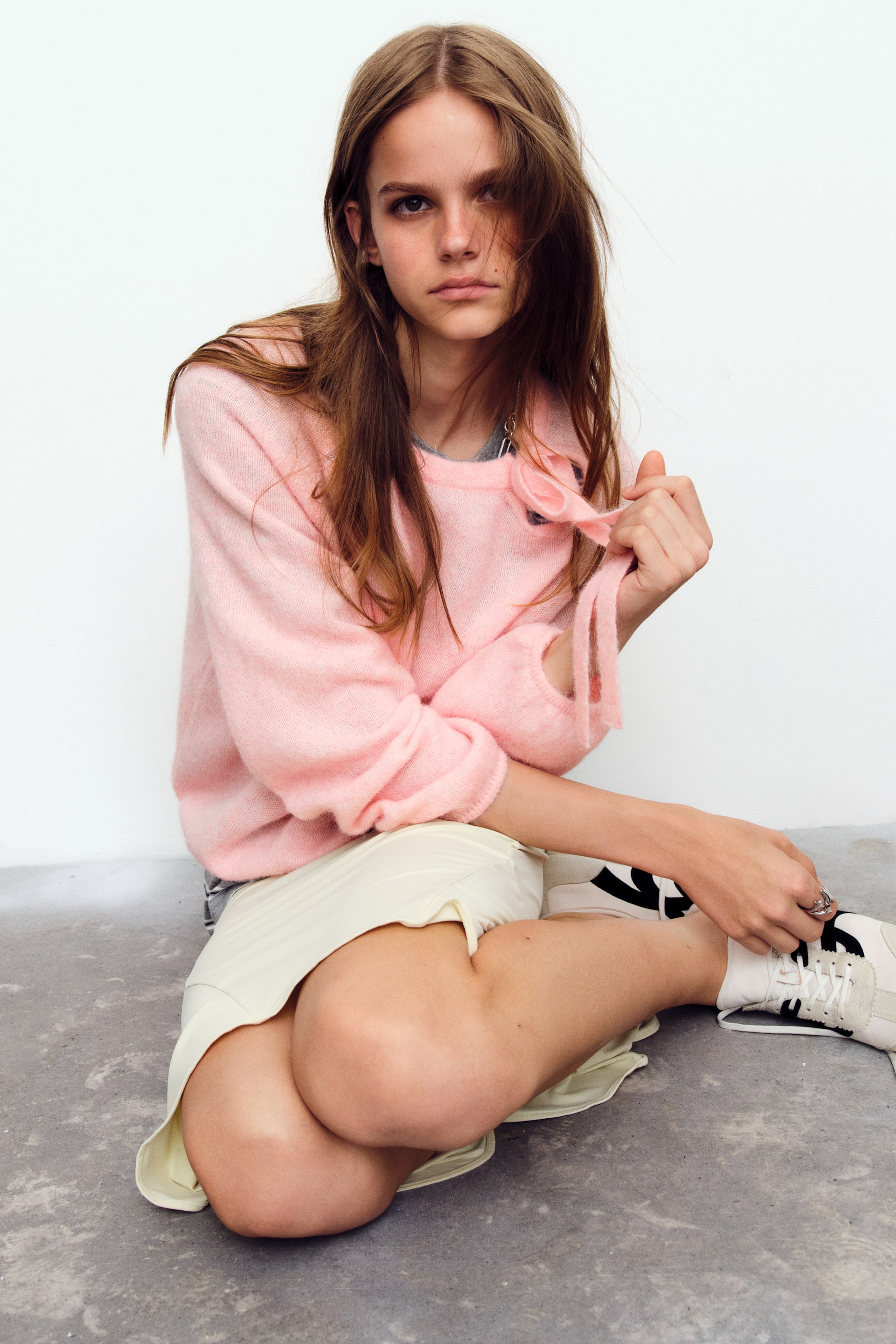 Ampliar la imagen: Una suéter rosa de corte holgado, confeccionada en un tejido de punto suave y cepillado, es llevada por la modelo, quien está sentada y sostiene el lazo de su cuello tipo moño. La suéter presenta mangas globo raglán con puños acanalados y se lleva sobre una prenda superior gris.