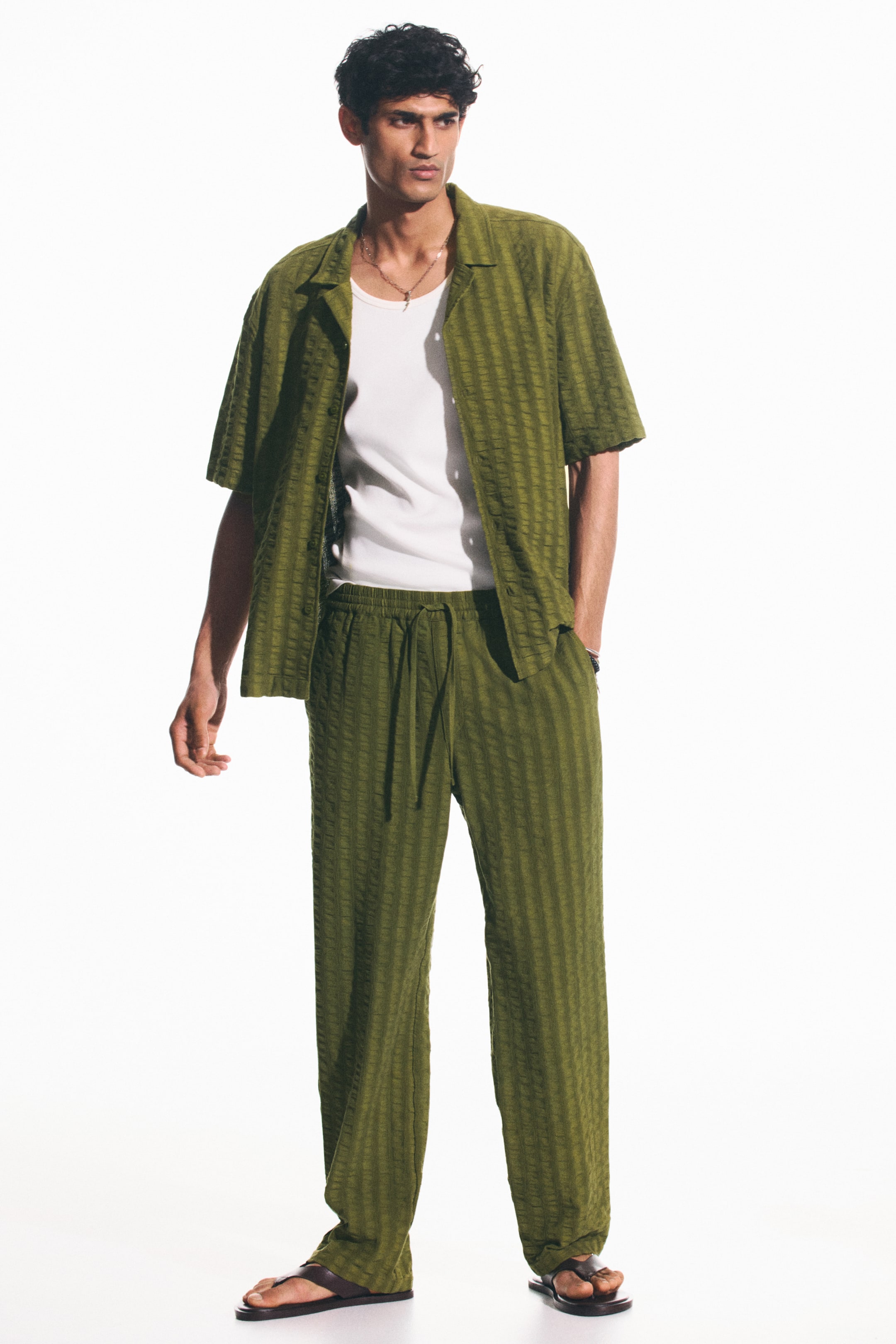 View larger image: "This olive green matching set showcases a short-sleeved, open-collar shirt styled open over a white undershirt, paired with relaxed-fit pants featuring a visible drawstring waist. Both pieces exhibit a subtle vertical textured stripe pattern, perfect for a laid-back yet stylish look."