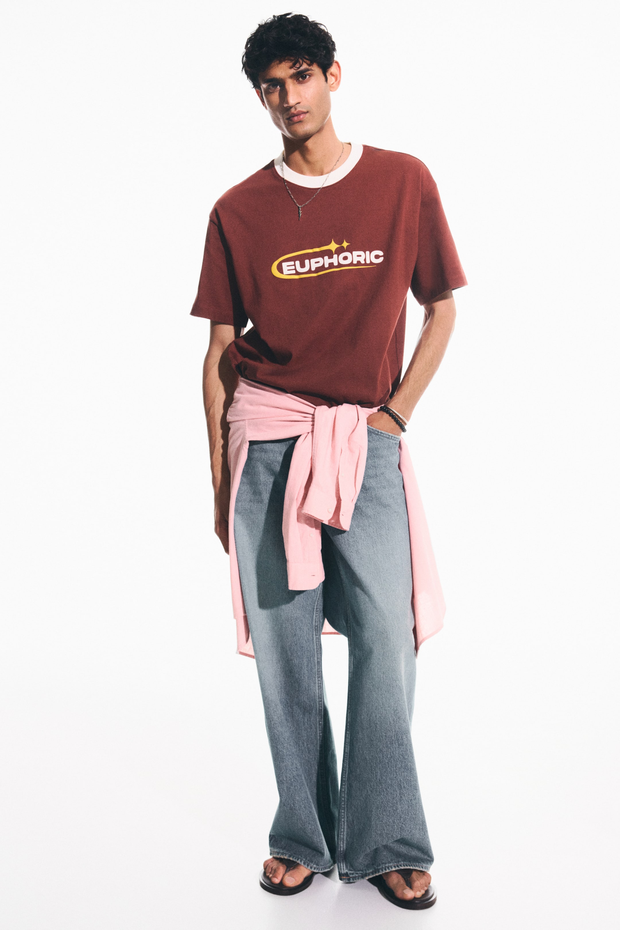 View larger image: A brown t-shirt with a white ribbed crew neck and a yellow 'EUPHORIC' graphic is styled with light blue wide-leg jeans and black flip-flops, complemented by a light pink shirt tied around the waist.