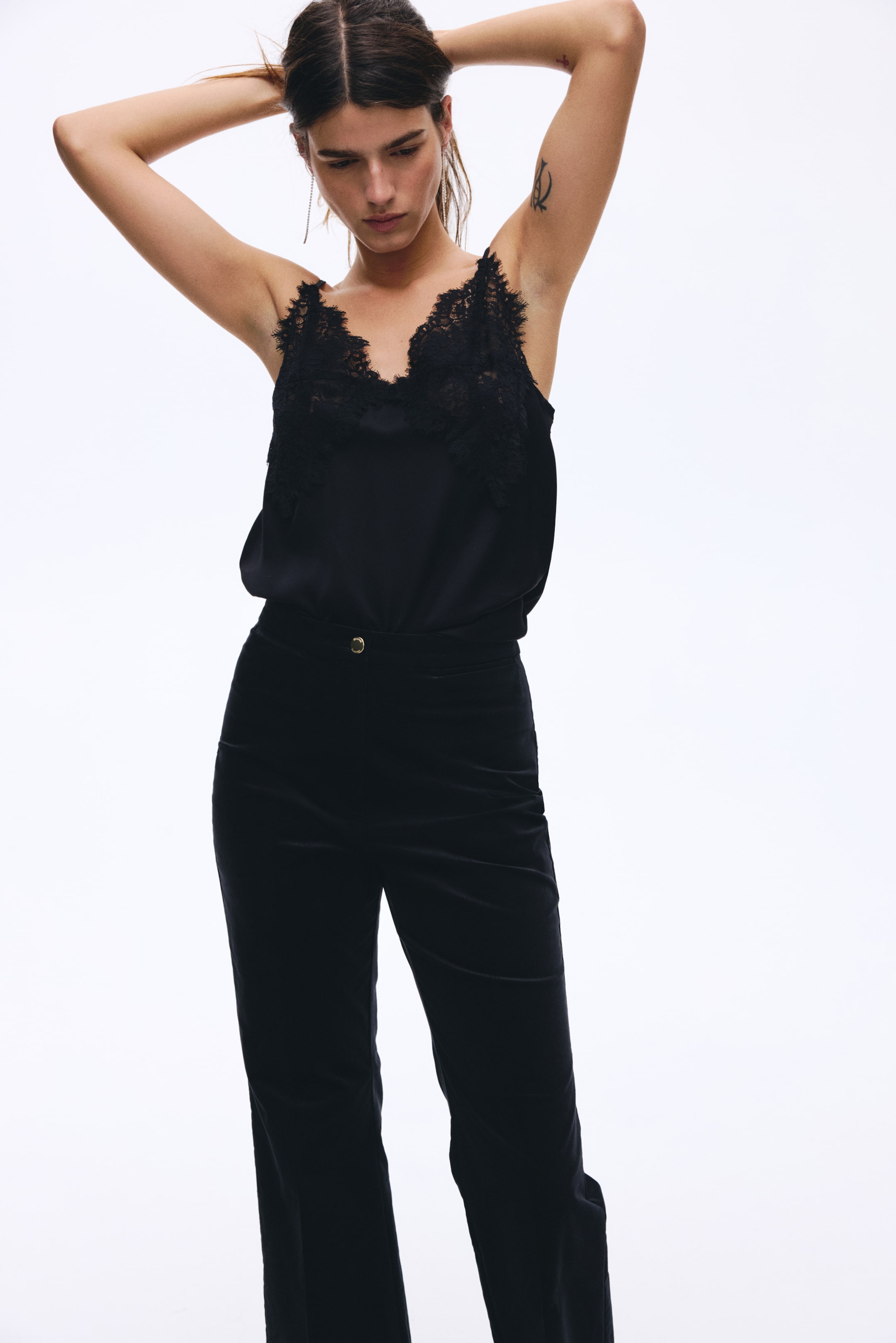 Ver imagen más grande: Una camiseta negra, con un escote en V y un ribete de encaje floral negro, se complementa con pantalones de terciopelo negro. Los pantalones de cintura alta presentan un cierre con un único botón dorado y un corte recto, todo ello mostrado desde un ángulo frontal.