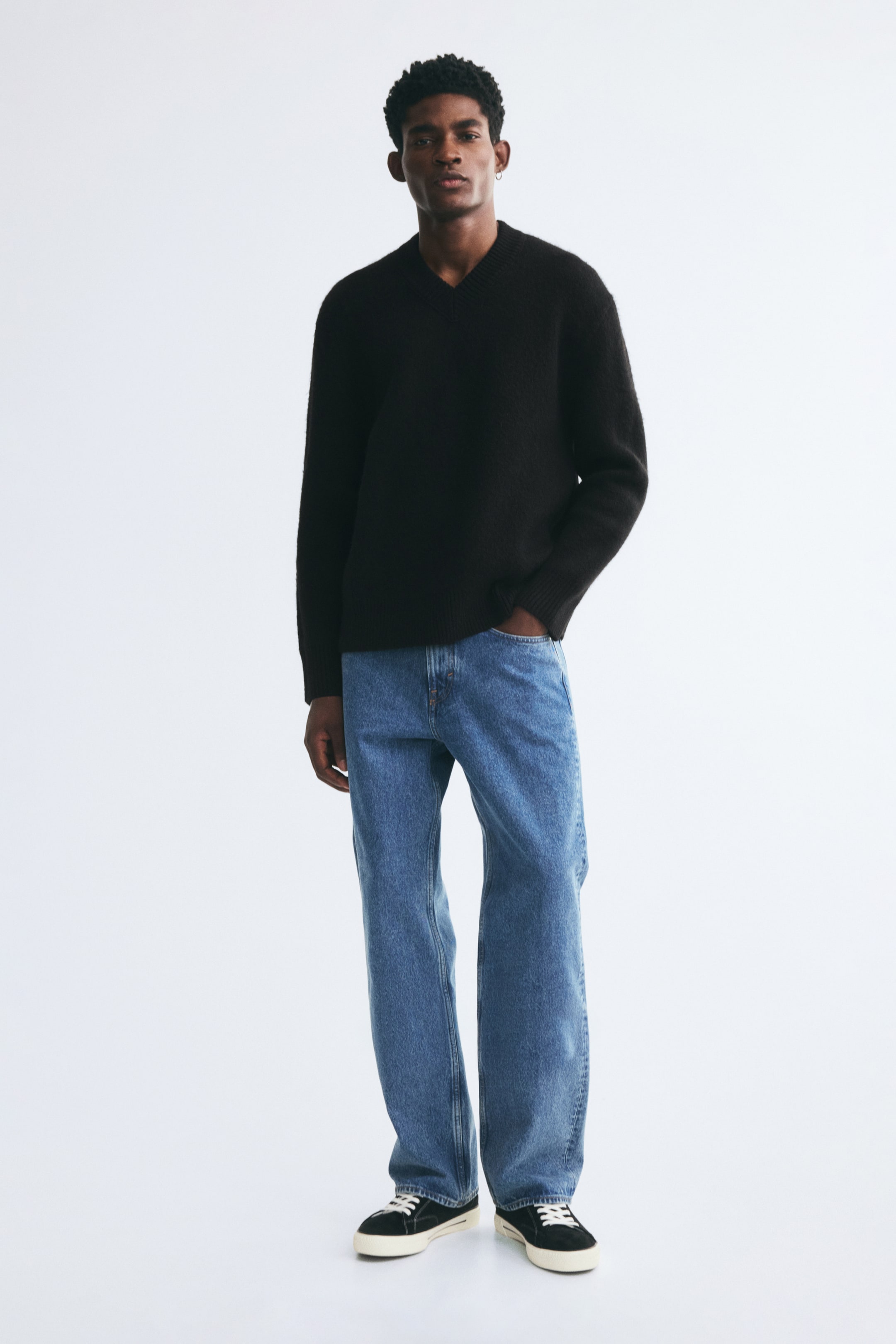 View larger image: A black knit sweater featuring a relaxed fit, a ribbed V-neck, and ribbed trim at the cuffs and hem is being worn by the model. The sweater is styled with blue jeans and black sneakers.