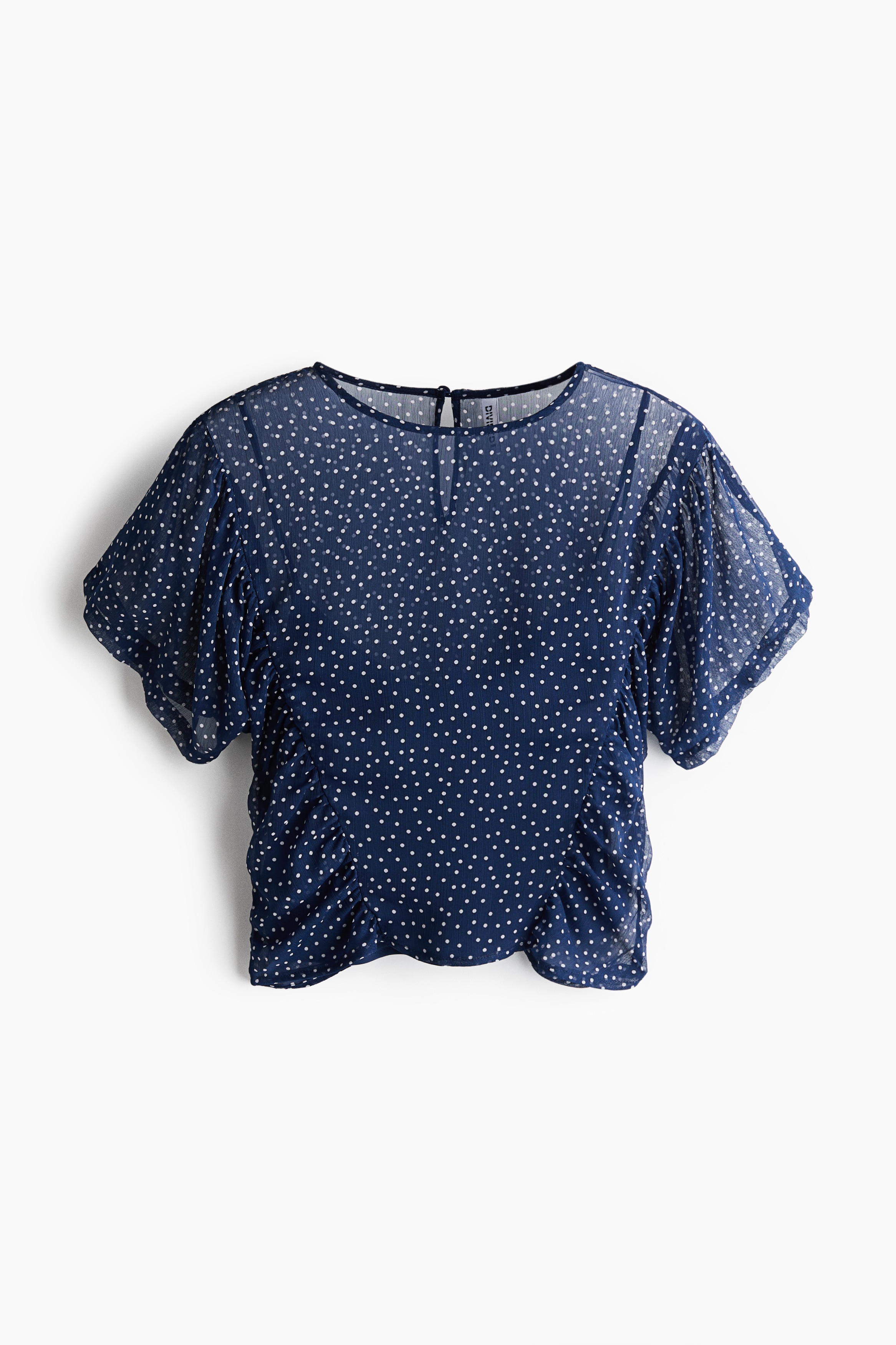 A sheer navy chiffon blouse with a white polka-dot pattern is being worn by the model, featuring a round neckline, short flutter sleeves, and a dark underlayer. The blouse is styled with dark, wide-leg denim jeans and light-coloured sneakers.