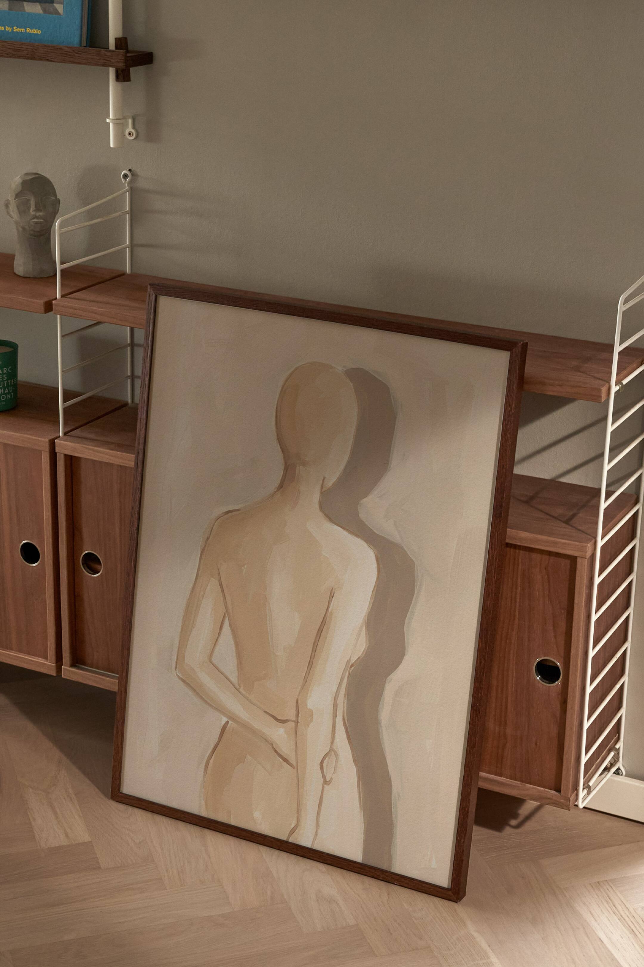 Agrandir l'image: Une affiche d'art orientée verticalement présentant une peinture minimaliste et stylisée du dos d’une silhouette, dans des tons neutres beige et brun clair, projetant une ombre douce vers la droite. L’œuvre est présentée dans un cadre en bois foncé et est légèrement inclinée.