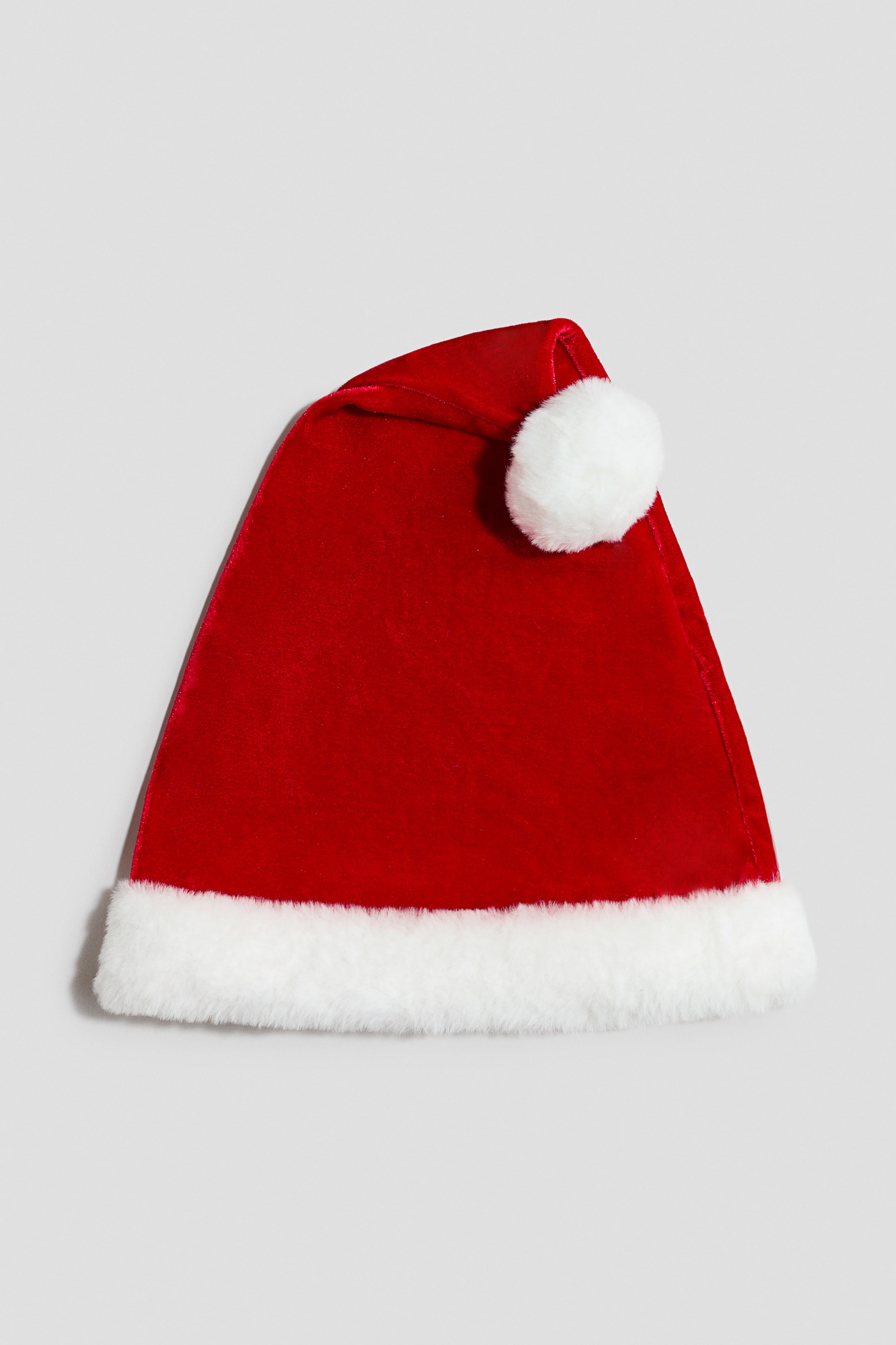 View larger image: A red velour Santa hat is lying flat in a front view, featuring a contrasting white fluffy trim at the hem and a fluffy white pompom on the folded tip.