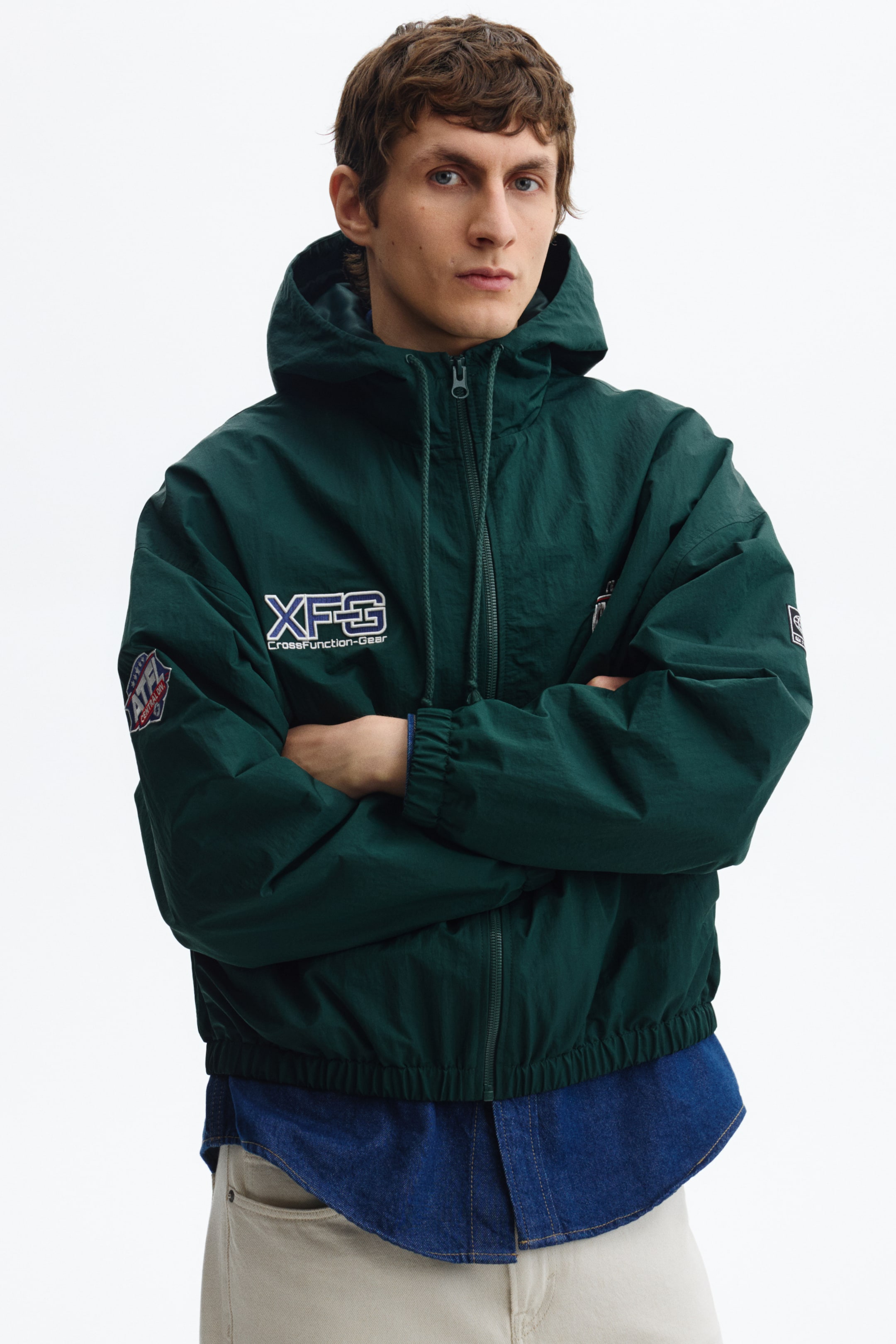 Vezi imaginea mai mare: O jachetă cu glugă, verde închis, este prezentată dintr-un unghi frontal. Aceasta are închidere cu fermoar pe întreaga lungime, glugă atașată cu șnururi, manșete elasticizate și tiv elastic. Jacheta afișează un logo alb și albastru „XFG CrossFunction-Gear” pe pieptul stâng și o emblemă în formă de scut „ATL ATHLETIC GEAR” pe mâneca stângă și este purtată peste o cămașă din denim albastră.