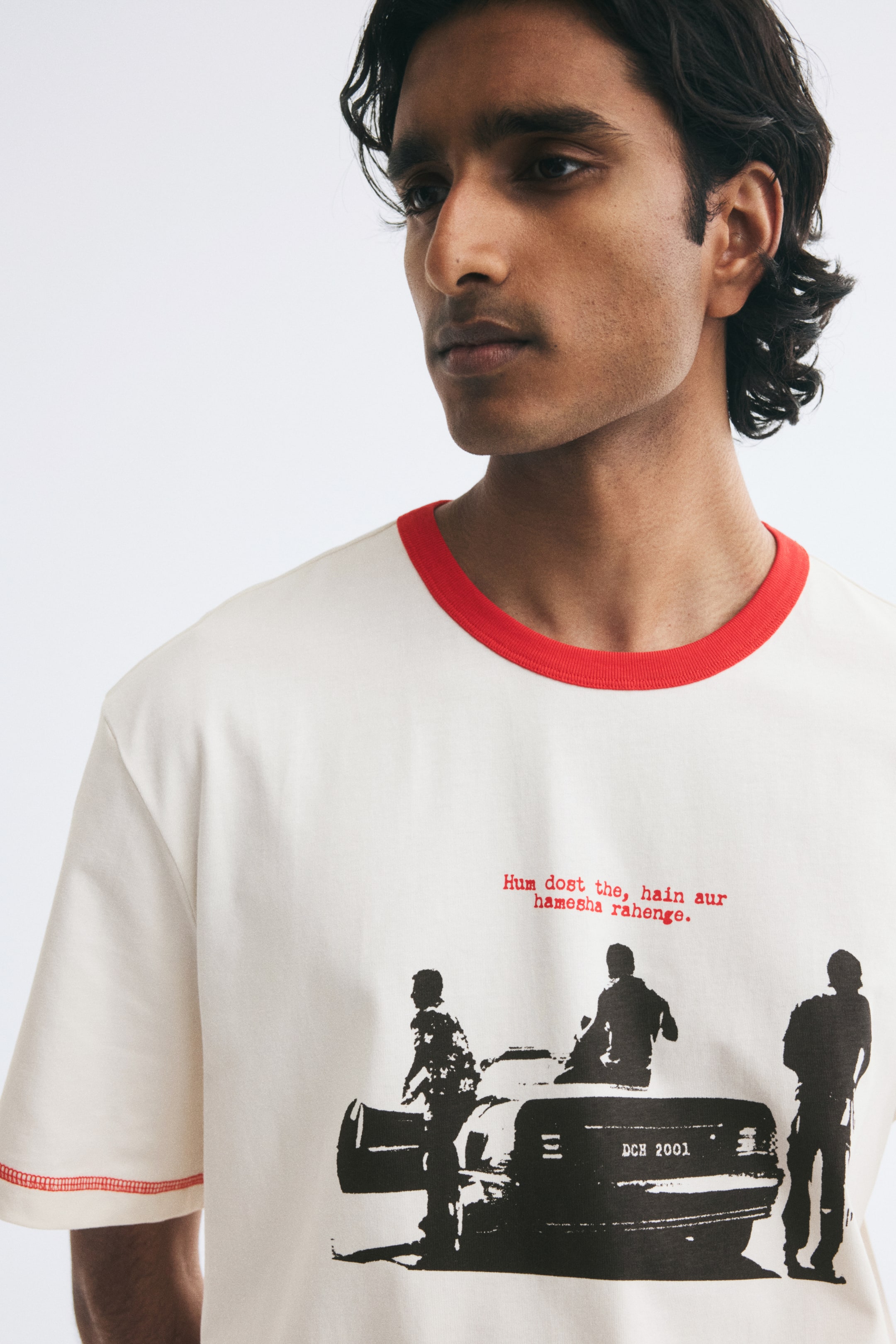 View larger image: A cream-coloured cotton jersey T-shirt is worn by the model, featuring a contrasting red ribbed neckline and a black printed graphic on the chest with red text. The short-sleeved tee also features red flatlock stitching on the sleeve cuffs.