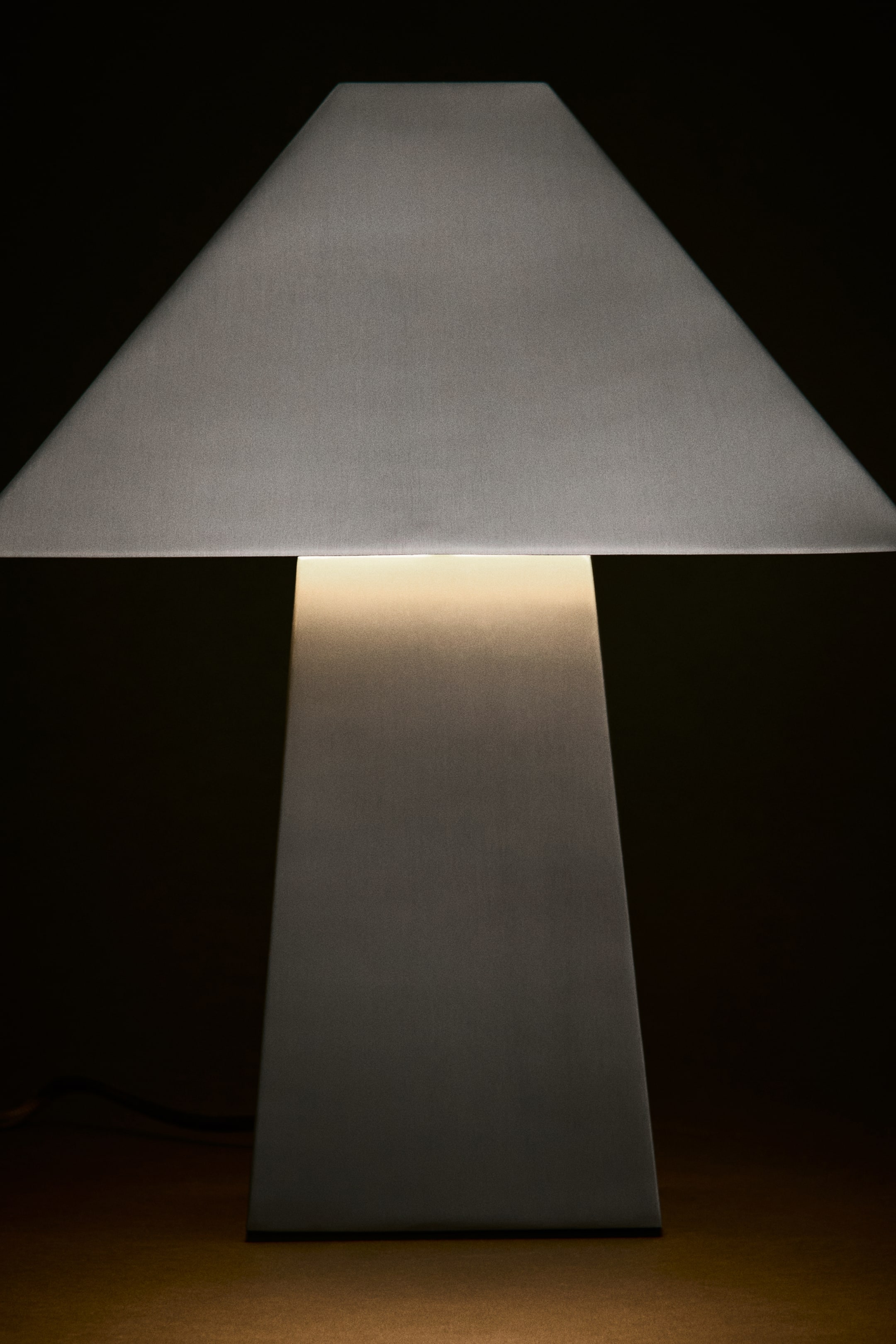 Zobacz większy obraz: Metalowa lampa stołowa widziana z przodu, o geometrycznym designie z trapezowym kloszem i zwężającą się prostokątną podstawą, stoi zapalona. Spod klosza emanuje ciepłe światło, rzucając miękkie odbicie na szczotkowaną metalową powierzchnię podstawy.
