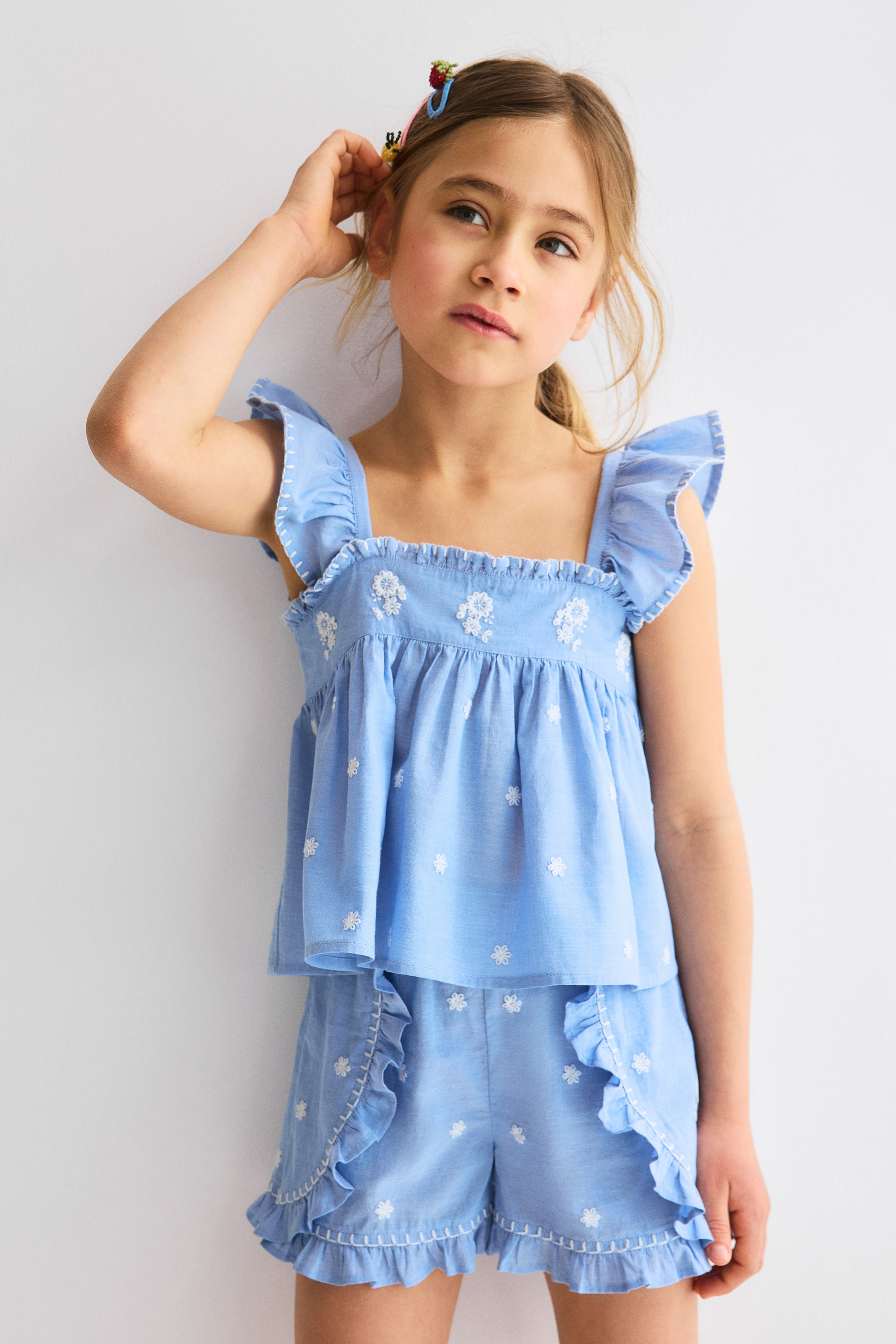 View larger image: The light blue two-piece set showcases a top with a square neckline, ruffled shoulder straps, and a gathered bodice. It is paired with matching shorts that feature ruffled side trim and a ruffled hem. Both pieces are adorned with white embroidered floral motifs and contrasting white stitching, adding a touch of elegance to the ensemble.