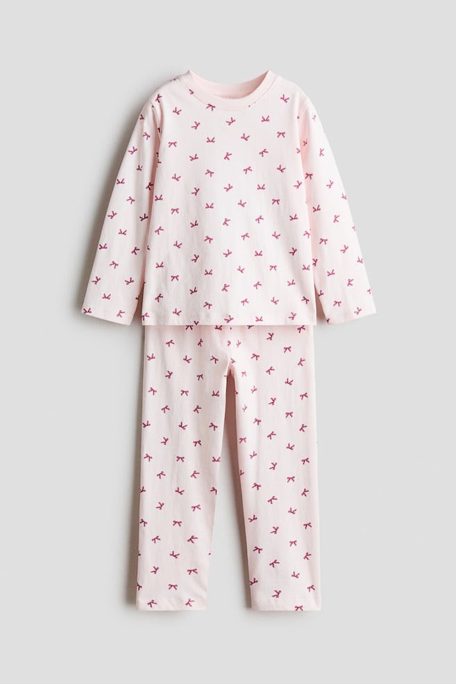 Ett ljusrosa pyjamasset i bomullsjersey visas framifrÃ¥n och bestÃ¥r av en lÃ¥ngÃ¤rmad topp och matchande byxor med ett Ã¶vergripande mÃ¶nster av smÃ¥ mÃ¶rkrosa rosetter. Toppen har en rund halsringning med smal kant, och byxorna har en rak benpassform. - 1
