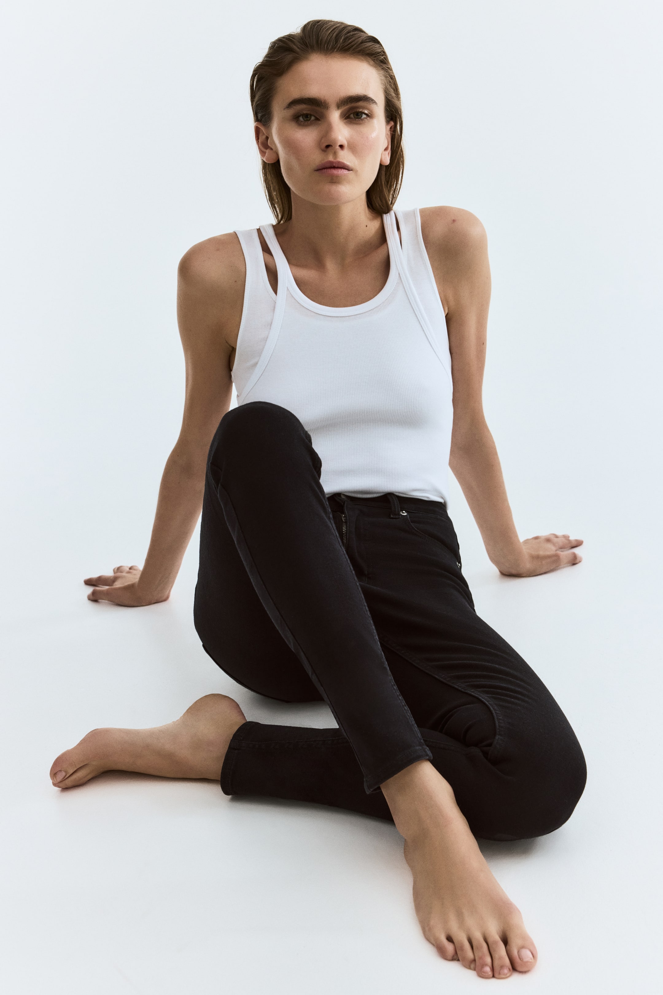 Größeres Bild anzeigen: Ein weißes geripptes Tanktop mit einem Rundhalsausschnitt und einem sichtbaren Doppelträger-Design auf der linken Schulter. Schwarze Skinny-Jeans mit einem Frontreißverschluss und Knopfverschluss, deren figurbetonte Struktur durch die Faltenbildung an den Knien beim Sitzen deutlich wird.