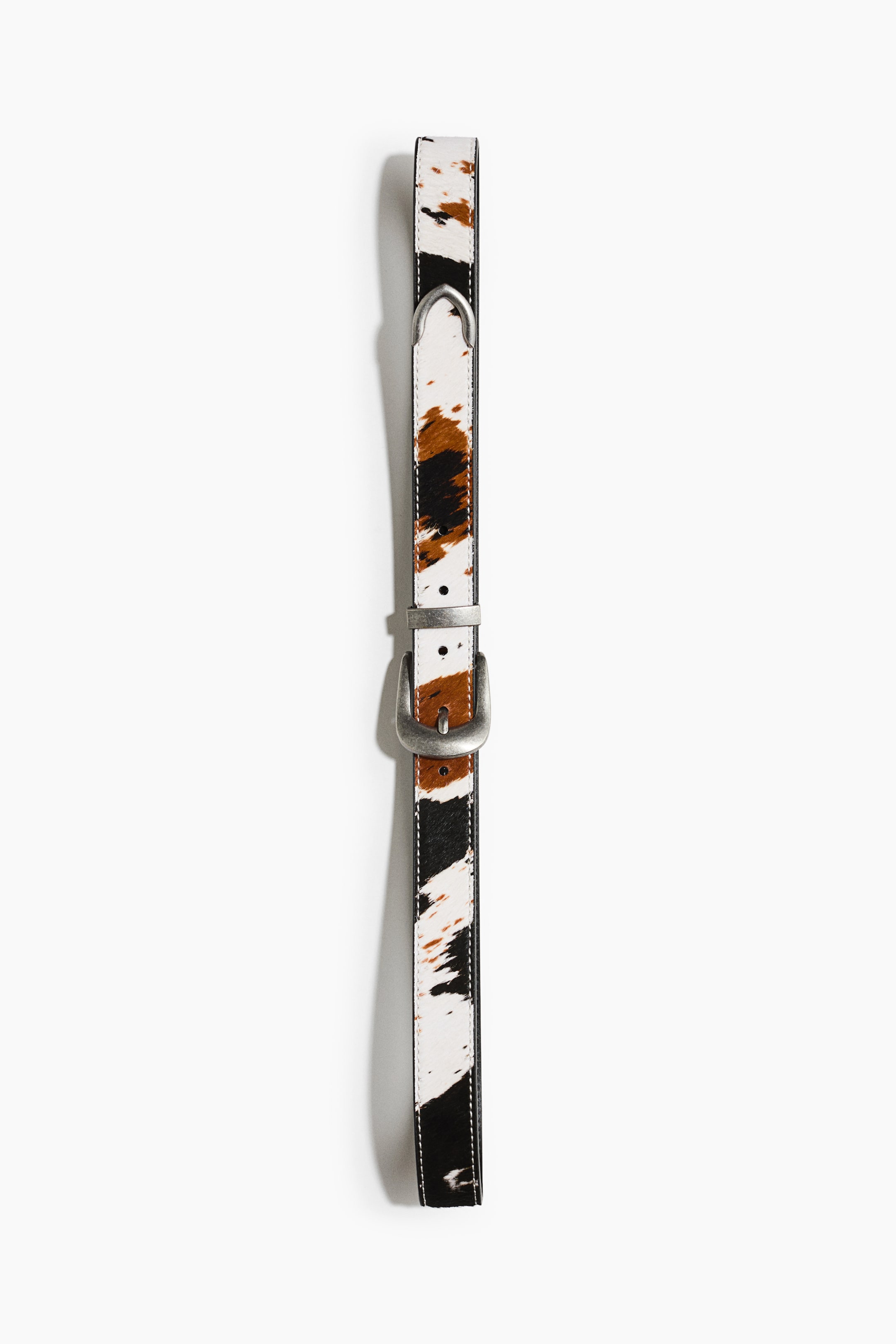 View larger image: A cowhide-patterned belt featuring a tricolor print in white, brown, and black is shown front-facing in a vertical position. It showcases a silver-tone western-style buckle, a matching metal keeper loop, and a pointed metal tip on the strap.
