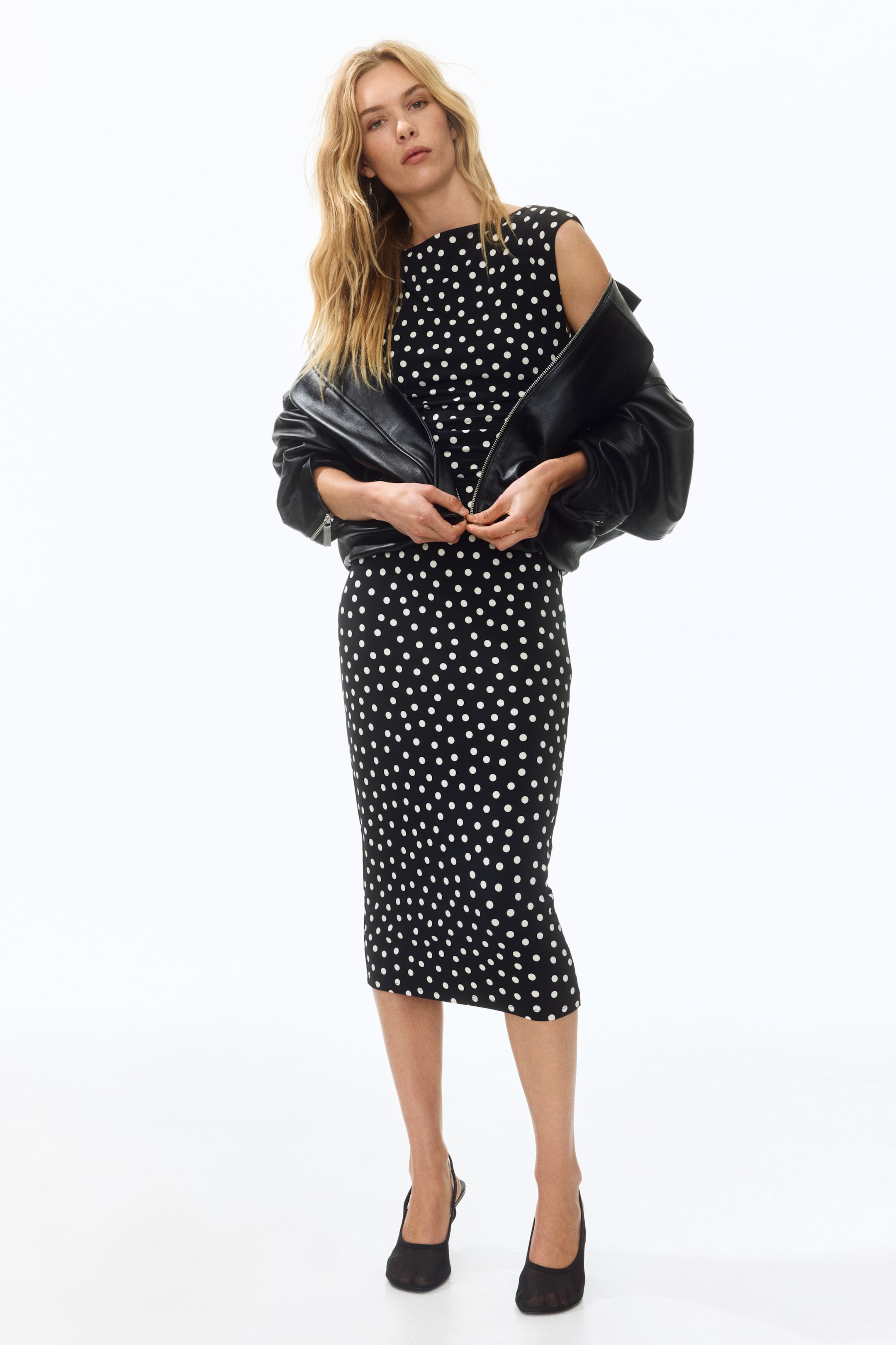 Voir une image plus grande: La robe midi noire présente un motif à pois blancs, un col bateau et un design sans manches. Elle offre une silhouette ajustée, avec une veste en cuir noir couvrant ses épaules.