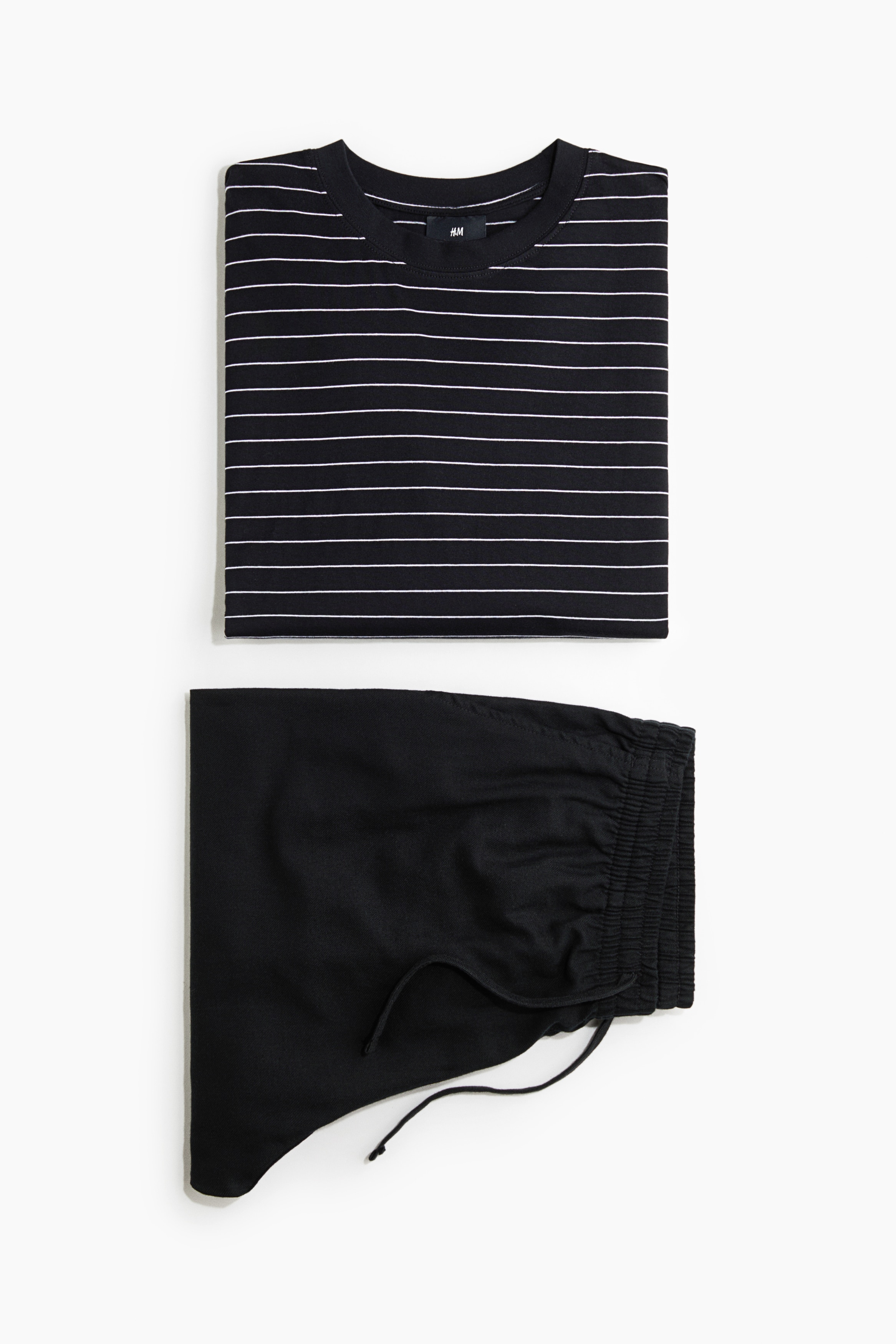 Een loungewear-set bestaande uit een zwart-wit horizontaal gestreepte jersey top met lange mouwen en een losse zwarte broek wordt gedragen door het model. De top heeft een ronde hals met ribboord en verlaagde schoudernaden, terwijl de broek is voorzien van een elastische tailleband met trekkoord.