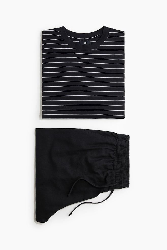 A folded two-piece pajama set is presented from the front, featuring a black crewneck t-shirt with thin, horizontal white stripes positioned above a pair of solid black shorts. The shorts are folded to display an elasticized waistband with a drawstring. - 1