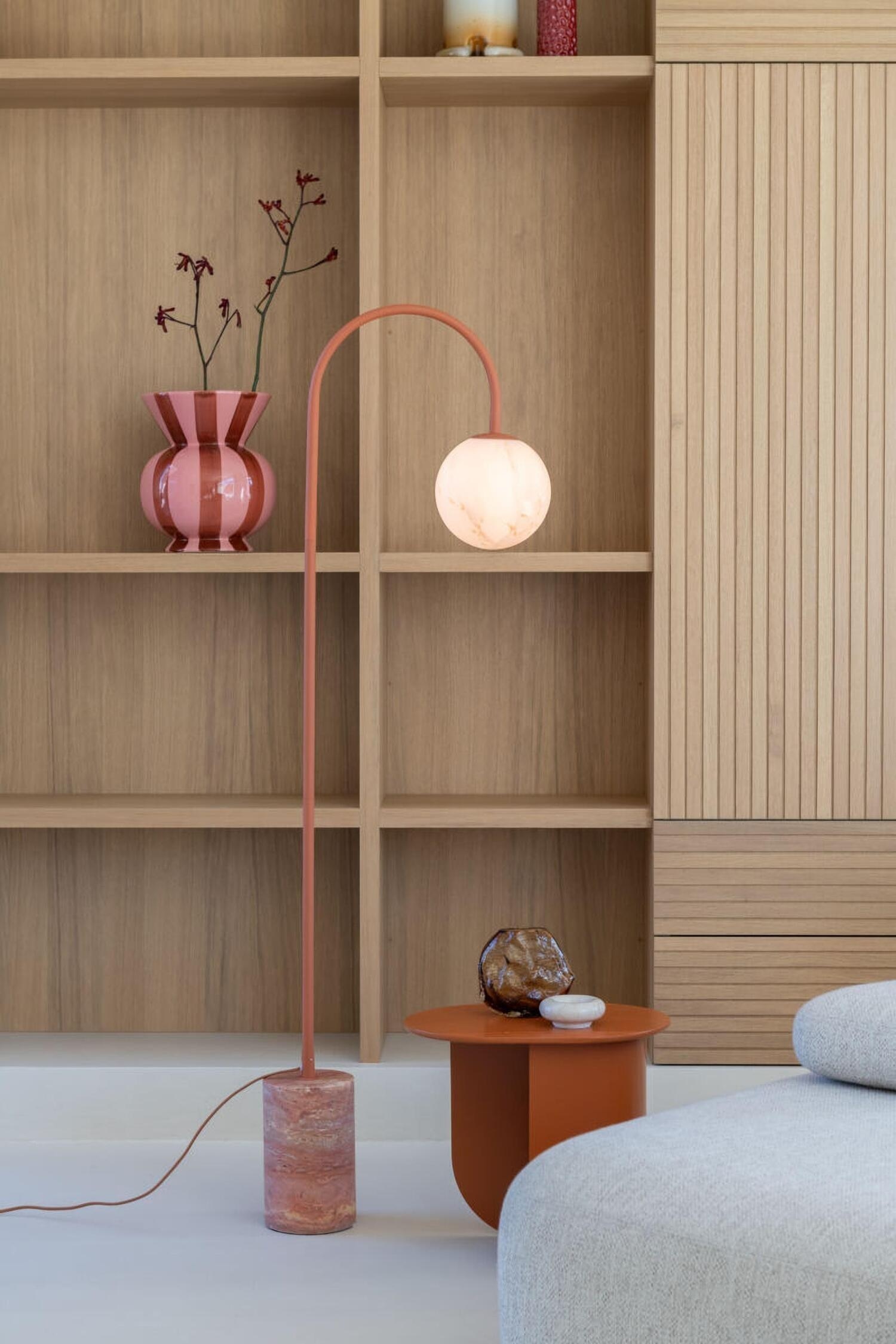 Un lampadaire est posÃ© sur une base cylindrique en pierre aux tons de terre superposÃ©s, avec une tige mÃ©tallique fine de couleur terracotta. La tige sâÃ©lÃ¨ve verticalement puis se courbe en une haute arche au sommet, dâoÃ¹ pend un abat-jour sphÃ©rique blanc translucide.