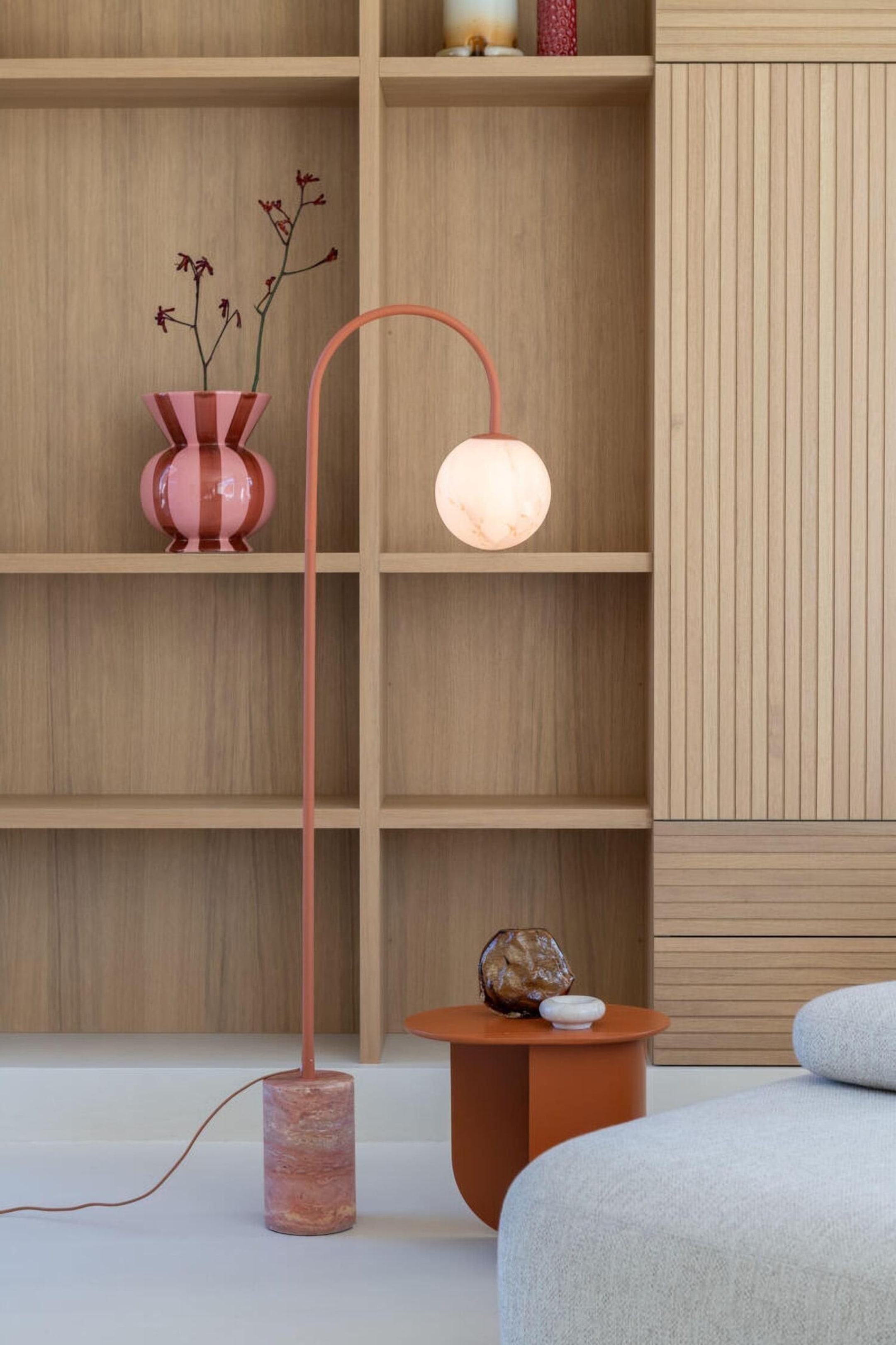 Agrandir l'image: Un lampadaire est posé sur une base cylindrique en marbre rose, avec une fine tige métallique en terre cuite, courbée en arche. La tige s’incurve vers le bas à son extrémité supérieure, suspendant un globe en verre blanc sphérique et lumineux.