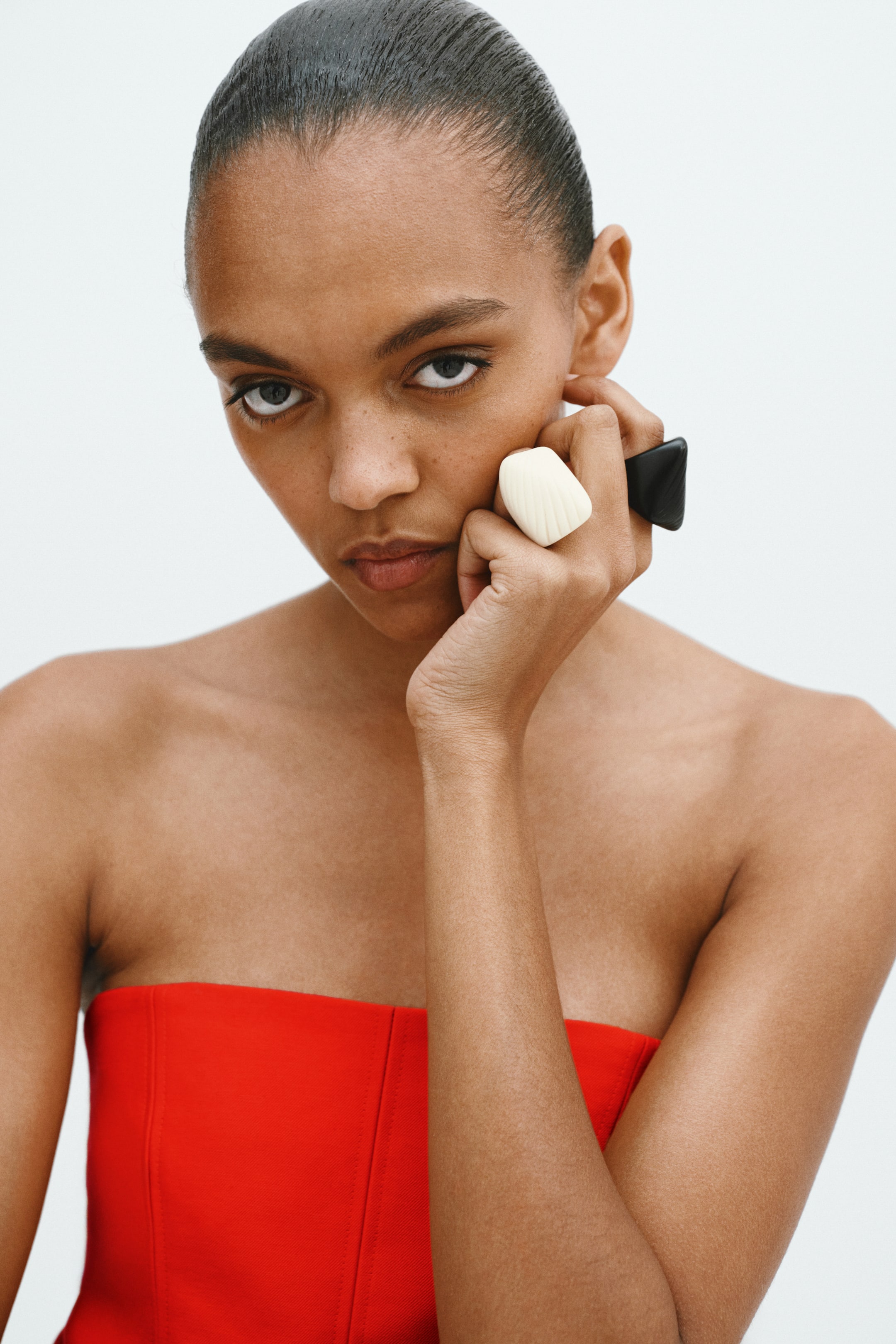 Ver imagem maior: Anéis de resina volumosos são usados nos dedos da modelo. A modelo está com a mão junto ao rosto, exibindo um anel off-white com acabamento esculpido e sulcado e um anel geométrico preto.