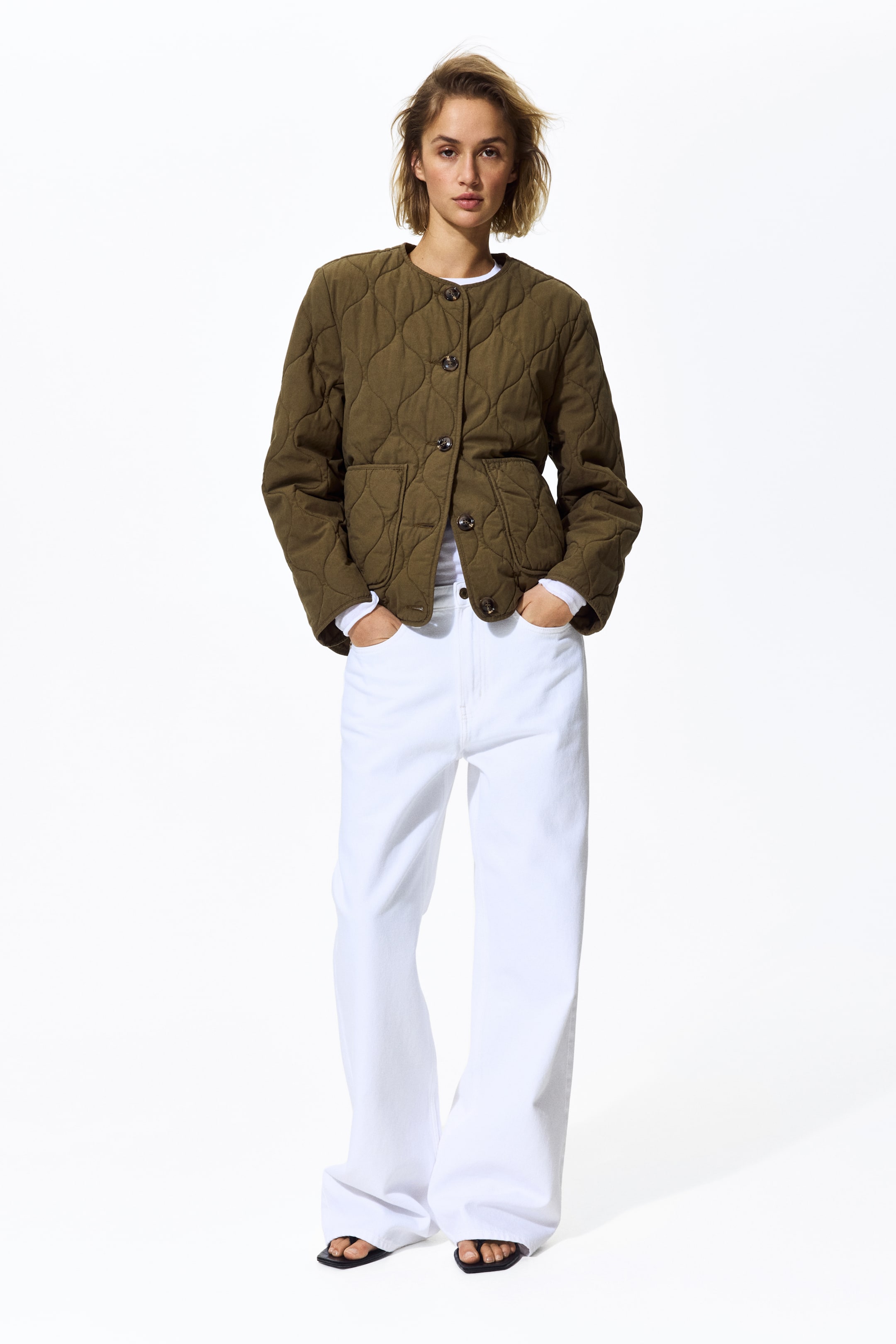 Ampliar la imagen: Una chaqueta acolchada verde oliva presenta un escote redondo sin cuello y un cierre frontal con botones oscuros. Está diseñada con un patrón de acolchado en forma de cebolla y dos bolsillos de parche en la parte inferior delantera, superpuesta sobre una camiseta blanca de manga larga.