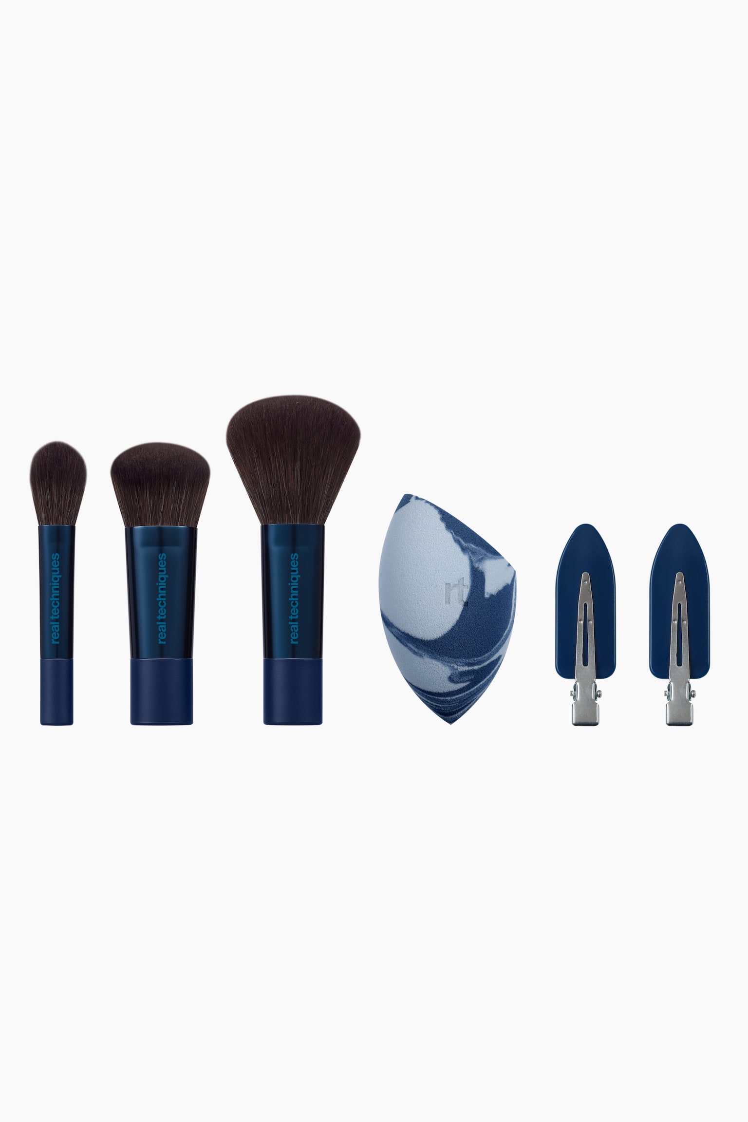 The Everyday Slay Brush & Sponge Set presenterar tre makeupborstar med metalliskt blå skaft och mörkbruna borst, en marmorerad blending-svamp i blått och vitt samt två blå creaseless-hårklämmor. Borstarna står upprätt i ökande storlek, medan svampen visar upp en platt vinklad kant och klämmorna har silverfärgade metallspännen.
