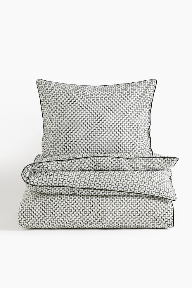 A bedding set consisting of an upright pillowcase and a folded duvet cover is displayed with a repeating black-and-white geometric grid pattern. Both items are facing forward and feature thin, dark green piped trim along the edges. - 1