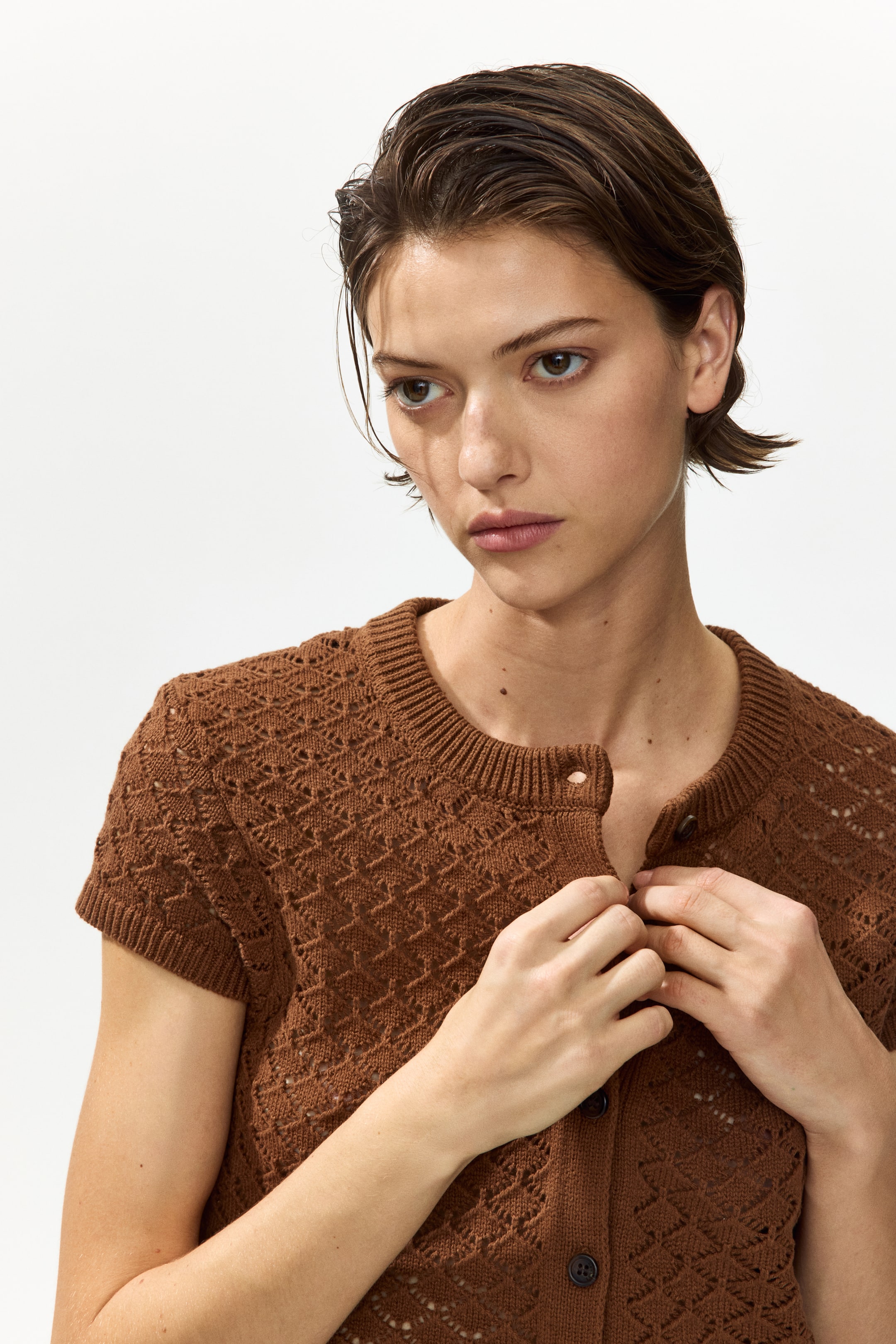 View larger image: The model is showcasing a short, fitted brown cardigan crafted in a pointelle knit. This piece sports a round neckline with ribbed trim, cap sleeves adorned with ribbed cuffs, and a button-down front which the model is in the process of fastening.