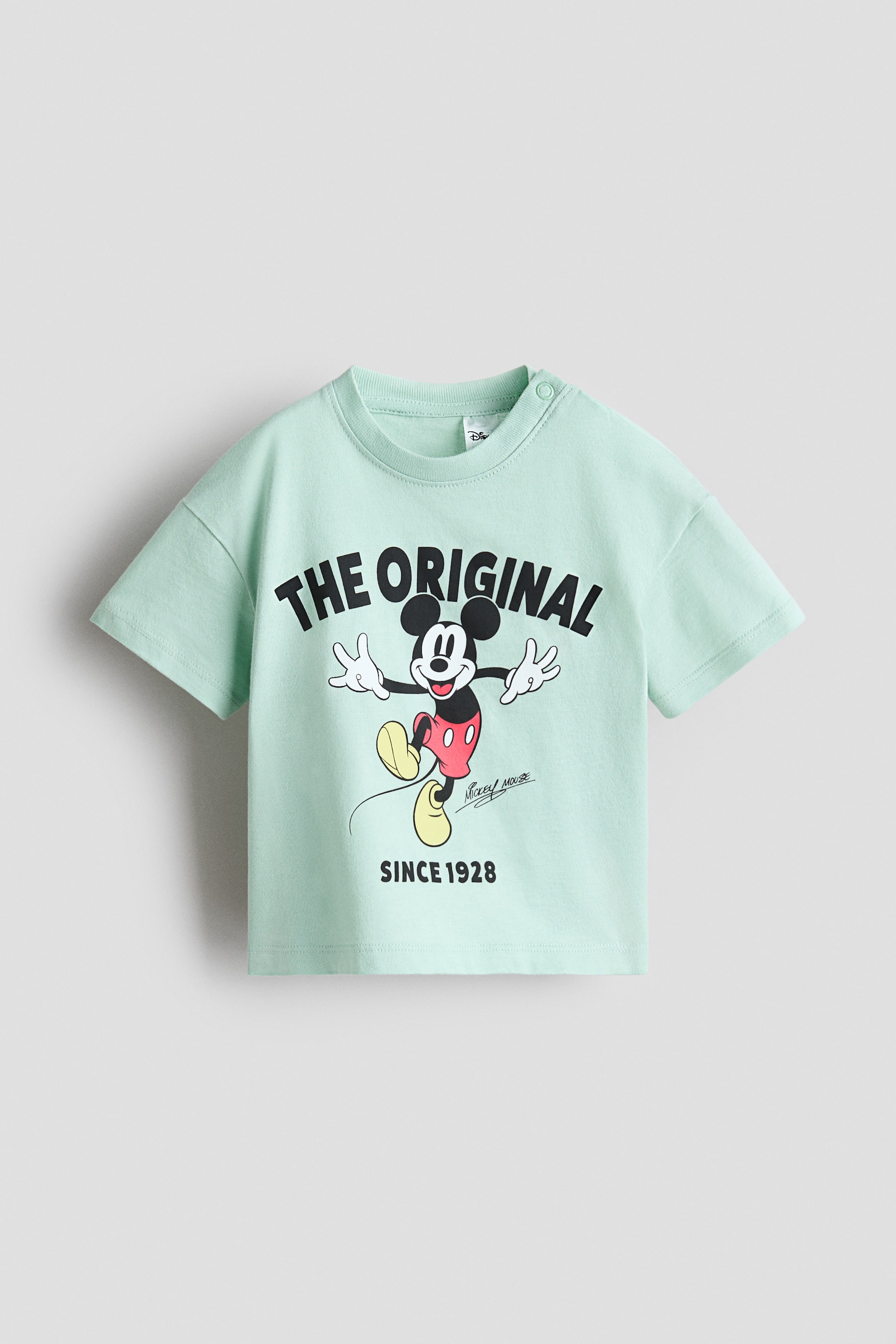 Voir une image plus grande: Un t-shirt à manches courtes vert pâle affiche un imprimé graphique de Mickey Mouse à l'avant, positionné entre le texte 'THE ORIGINAL' au-dessus et le texte 'SINCE 1928' en dessous. Il présente un col rond côtelé et deux boutons-pression sur l'épaule droite.
