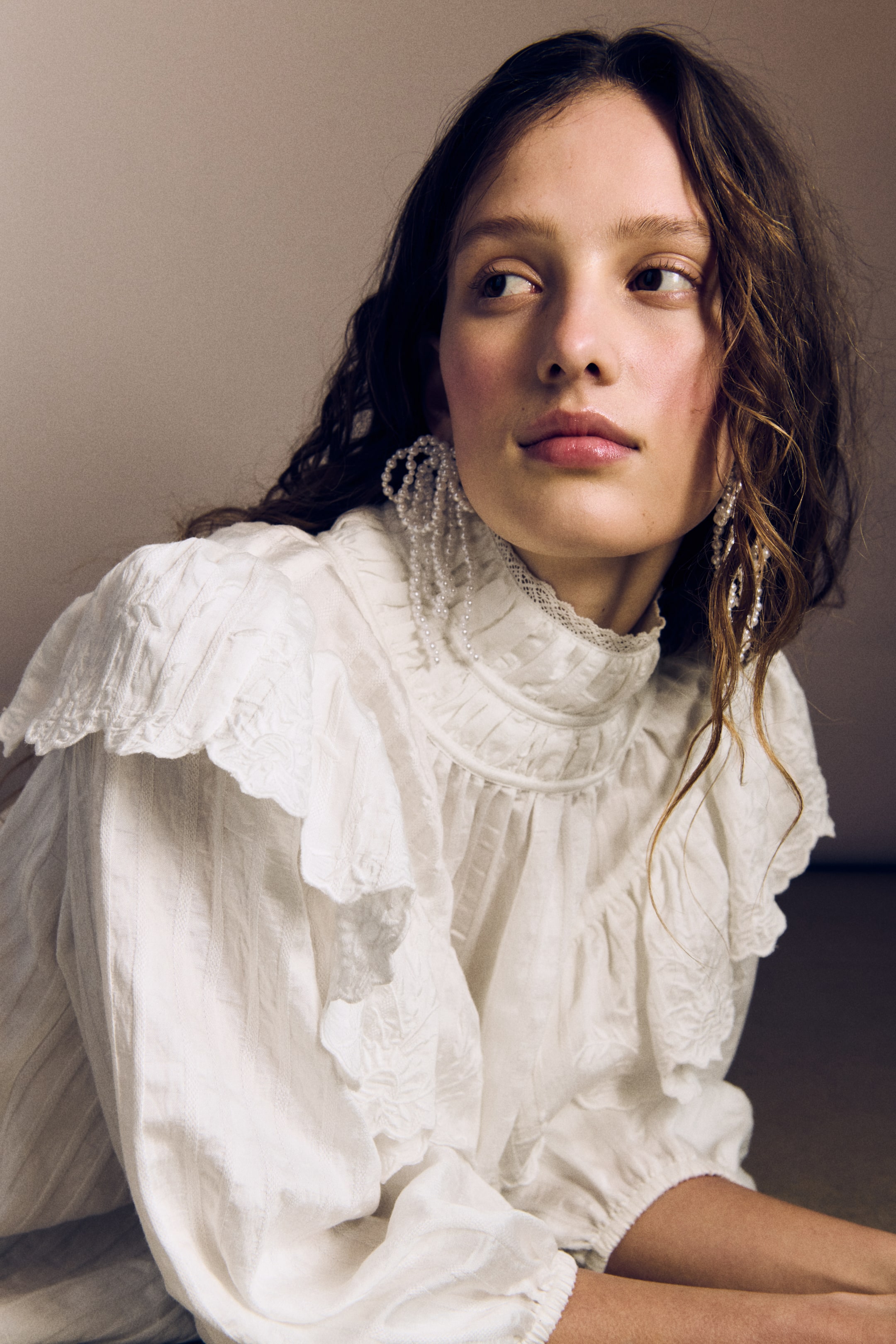 Agrandir l'image: Une blouse blanche à tissage texturé est portée par le mannequin, mettant en valeur un large volant brodé sur les épaules, un col roulé froncé orné de dentelle, et des manches ballon 3/4 avec poignets élastiqués.